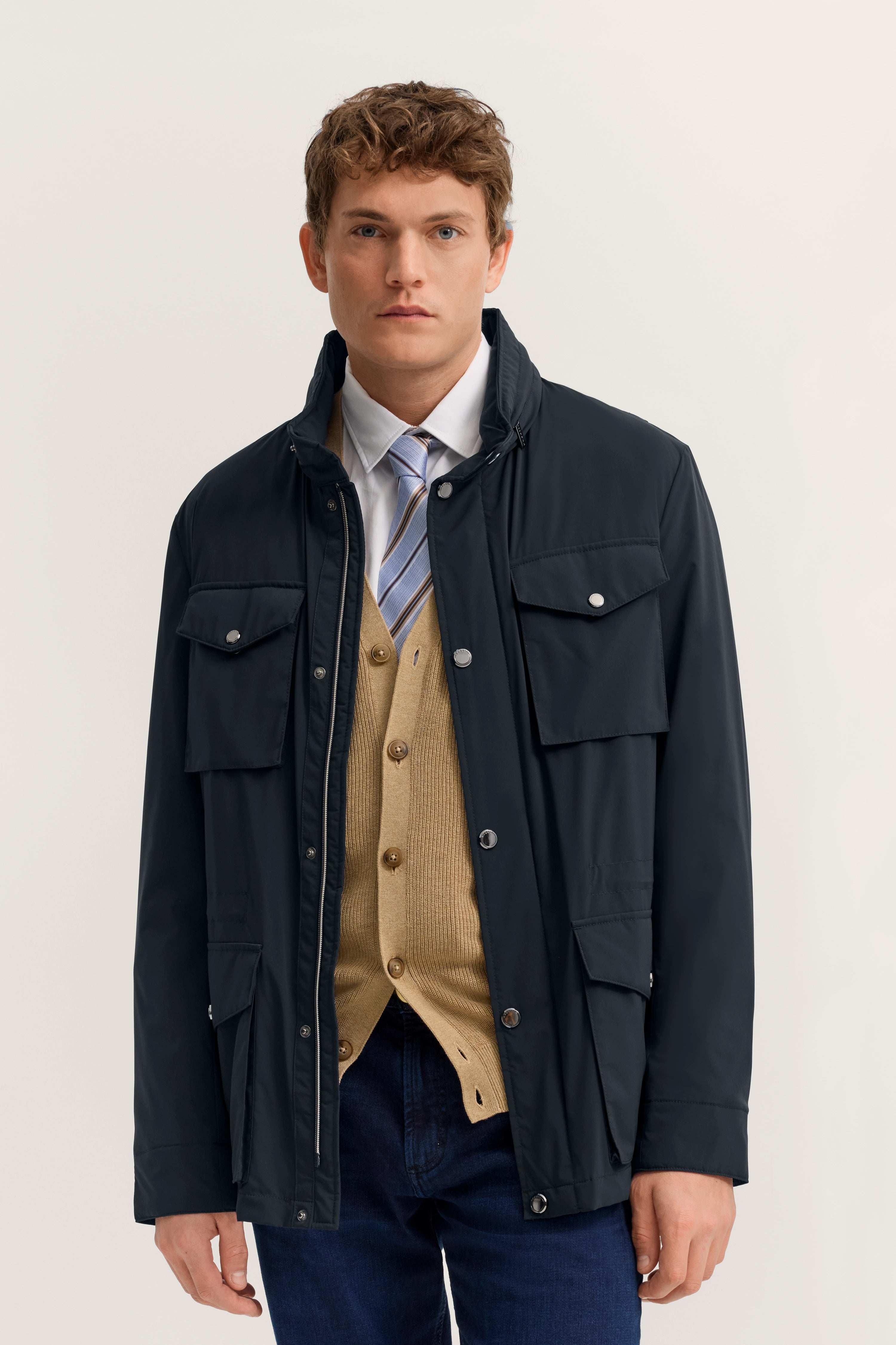 bugatti Model in dunkelblauer Jacke, beige Strickweste, weißem Hemd, gemusterter Krawatte, lässig stehend.