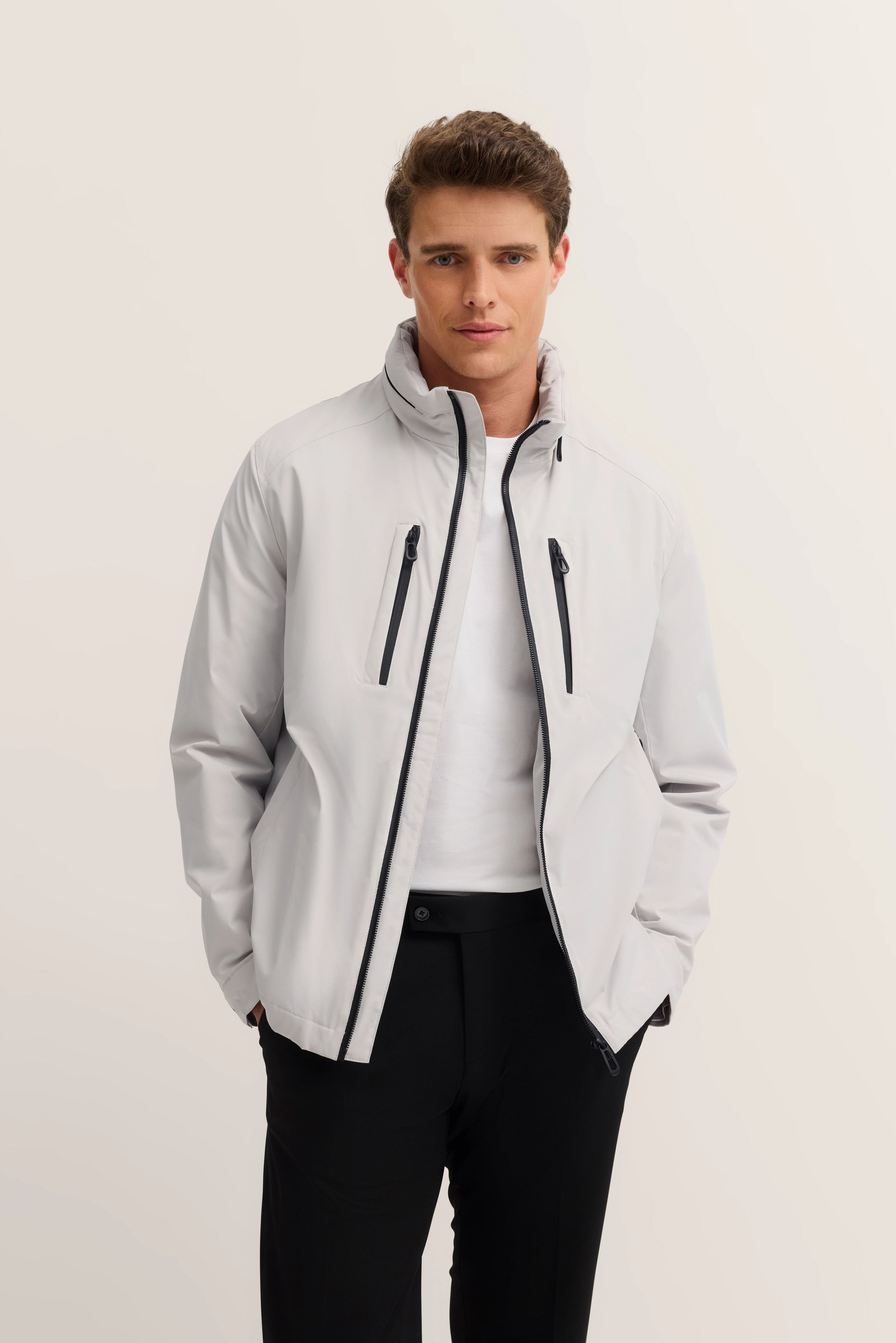 Model trägt graue bugatti Jacke mit schwarzem Reißverschluss, weißes Shirt, schwarze Hose, entspannte Pose.