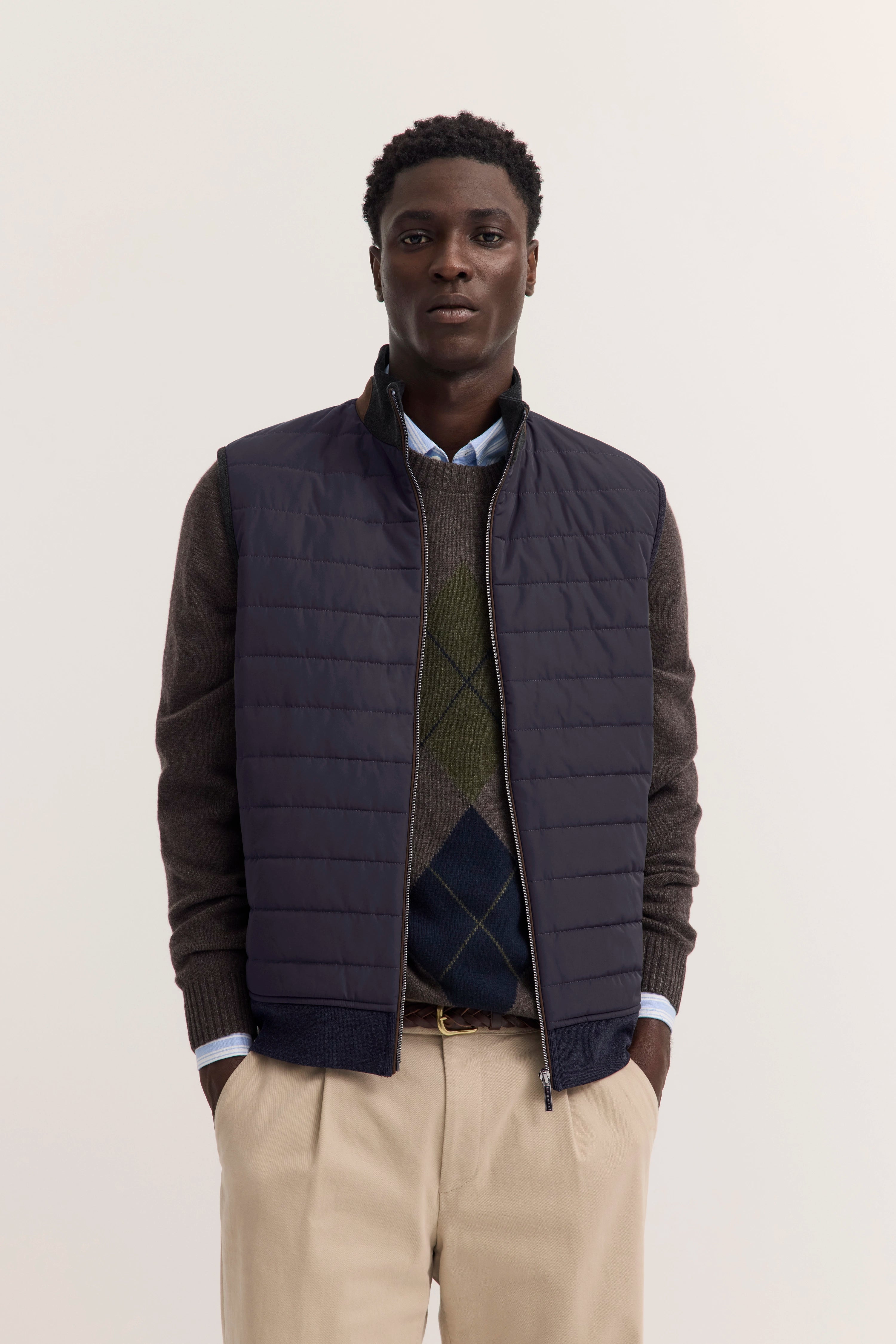 Bugatti Outfit: Dunkelblaue Weste, Rautenmuster-Pullover, hellblaue Hemd, beige Hose. Model stehend, Hände in Taschen.