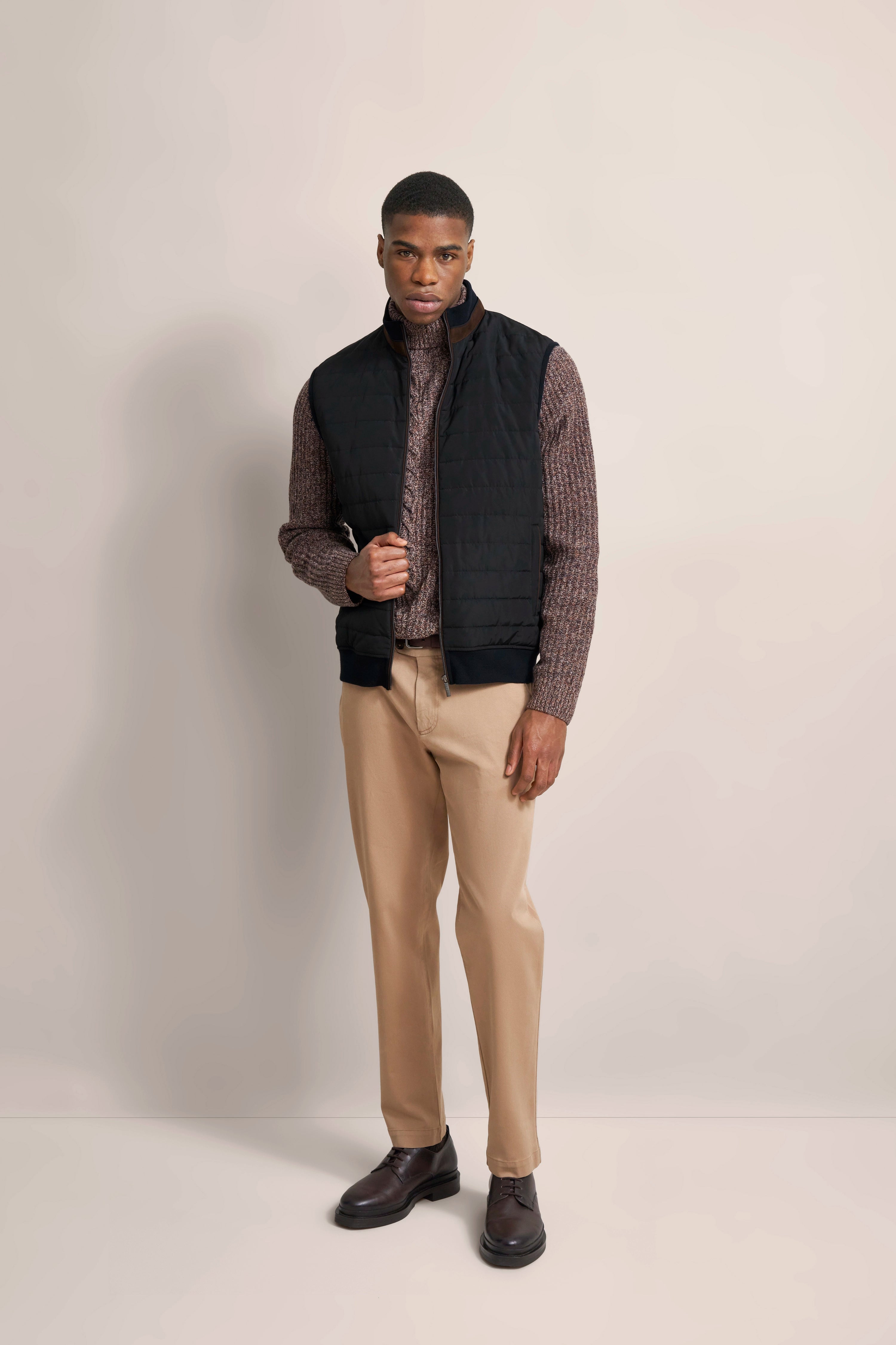 bugatti Outfit: Modell in schwarzer Weste, braunem Strickpullover, beigen Hosen, braunen Schuhen, lässige Pose.
