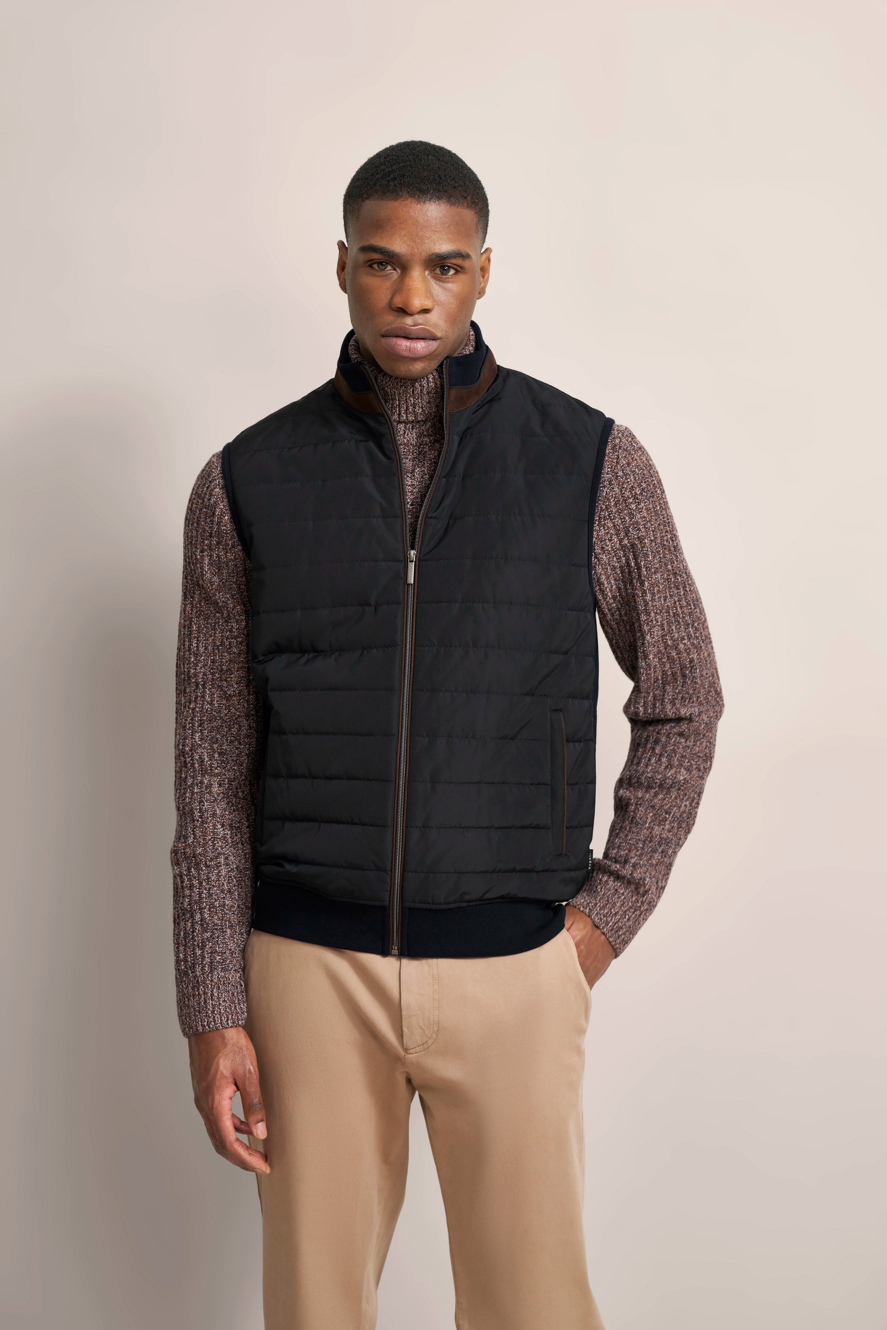 Model in schwarzer Steppweste, braunem Strickpullover, hellbeige Hose. Stehposenbild.