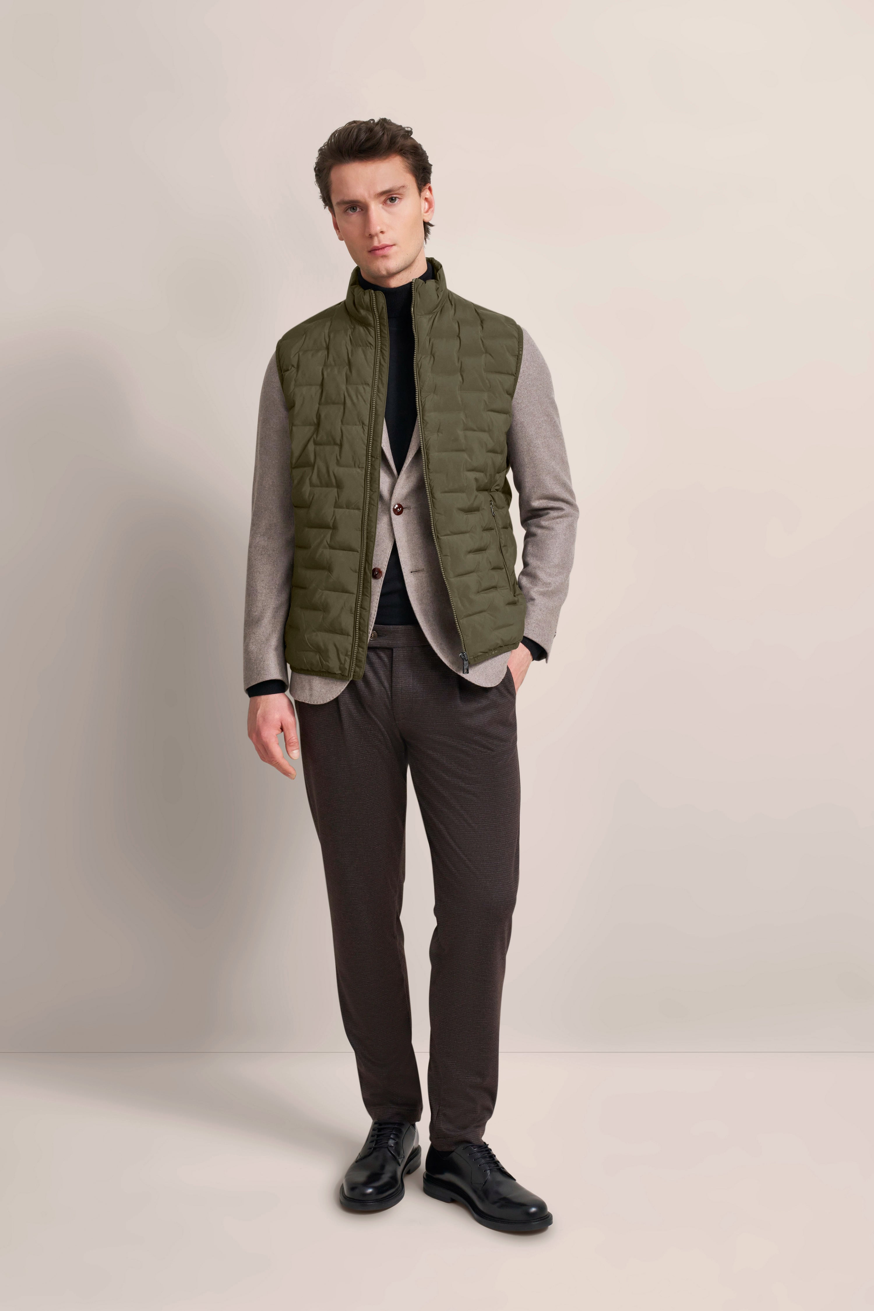 Model in olivgrüner Steppweste, grauem Sakko und schwarzem Rollkragen, braune Hose, schwarze Schuhe.