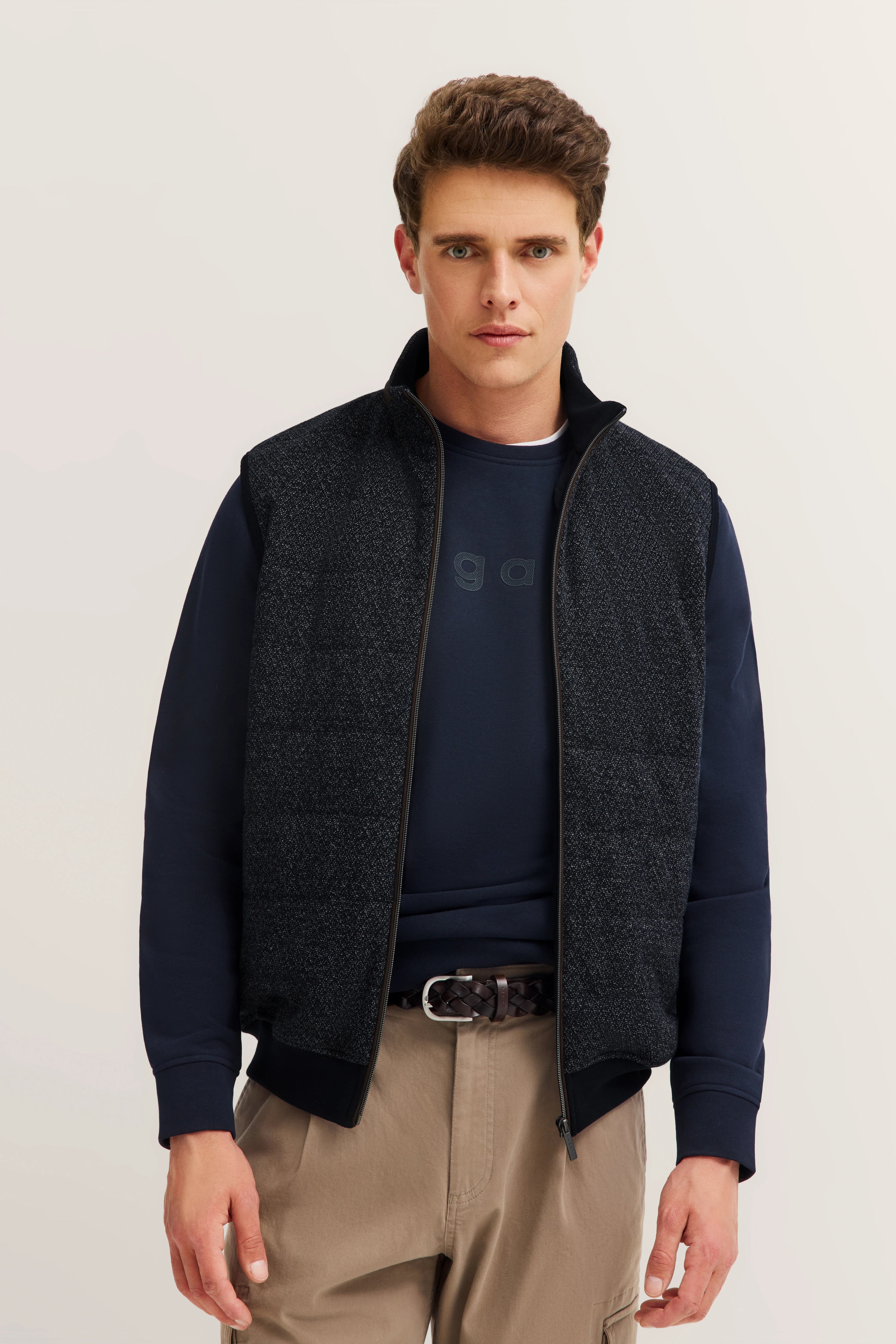 Model trägt bugatti Outfit: Dunkelblauer Pullover, graue Weste, beige Hose. Lässig stehendes Posing.