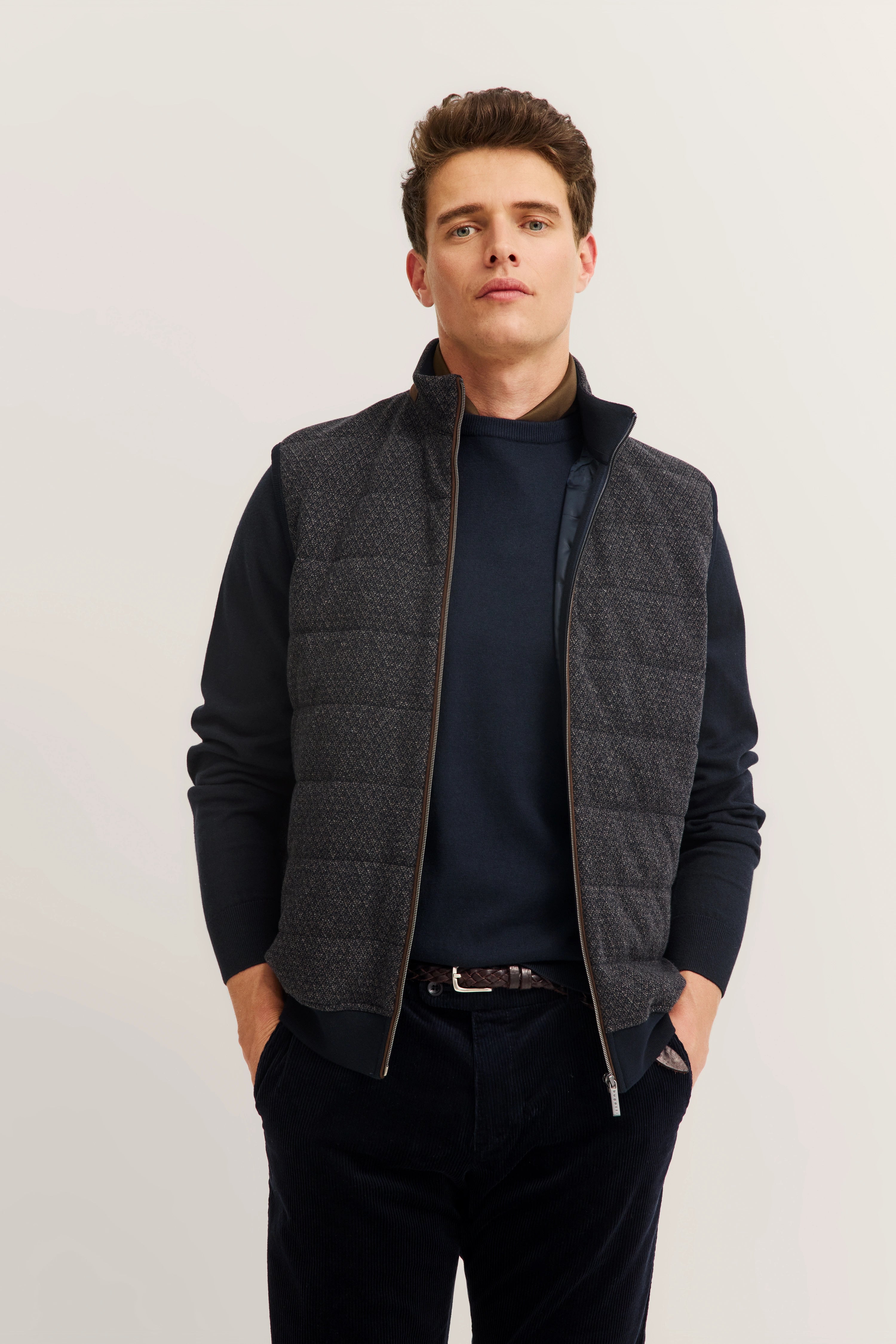 Bugatti Modelbild: Dunkelblauer Pullover, graue gesteppte Weste, entspannte Pose, Hände in Hosentaschen.