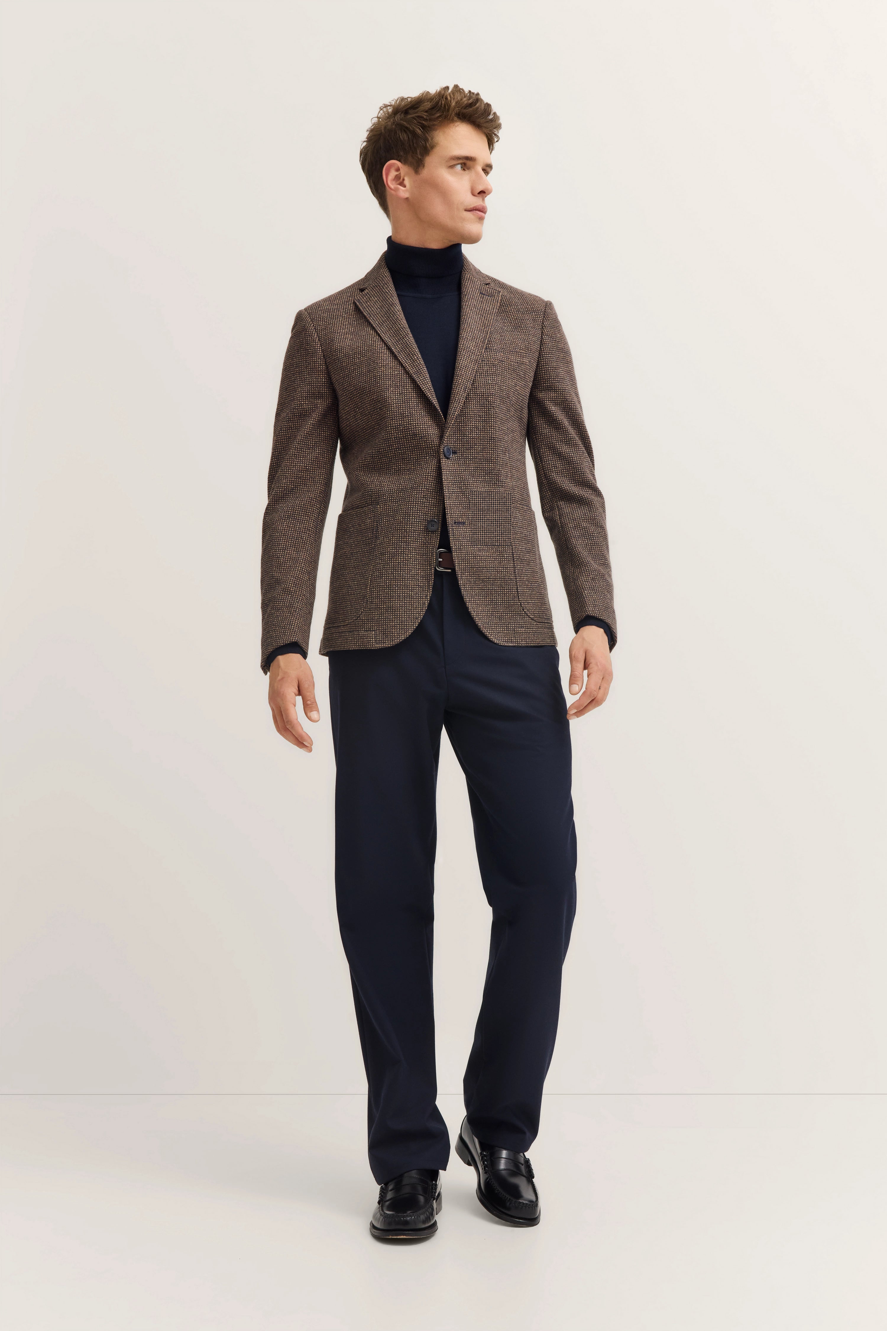 Model in brown blazer, dark blue pants, black turtleneck, side pose.