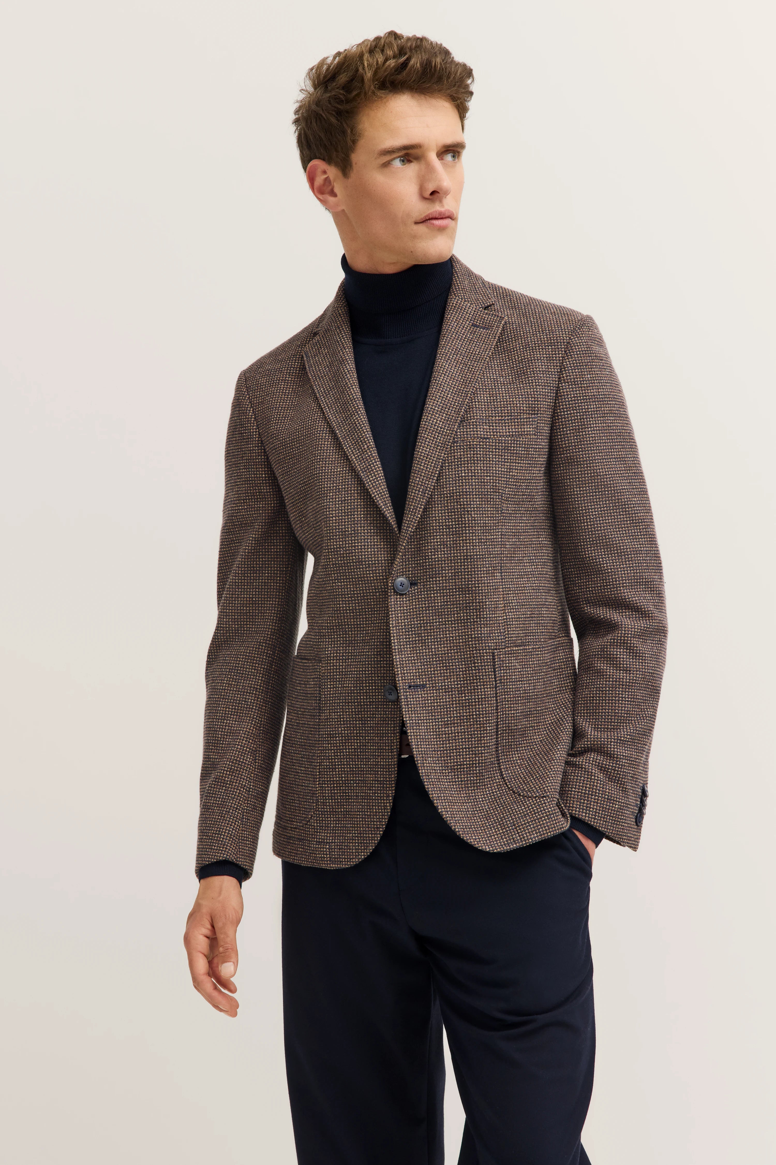 Model in brown bugatti blazer with pattern, black turtleneck, blue pants, posing casually.