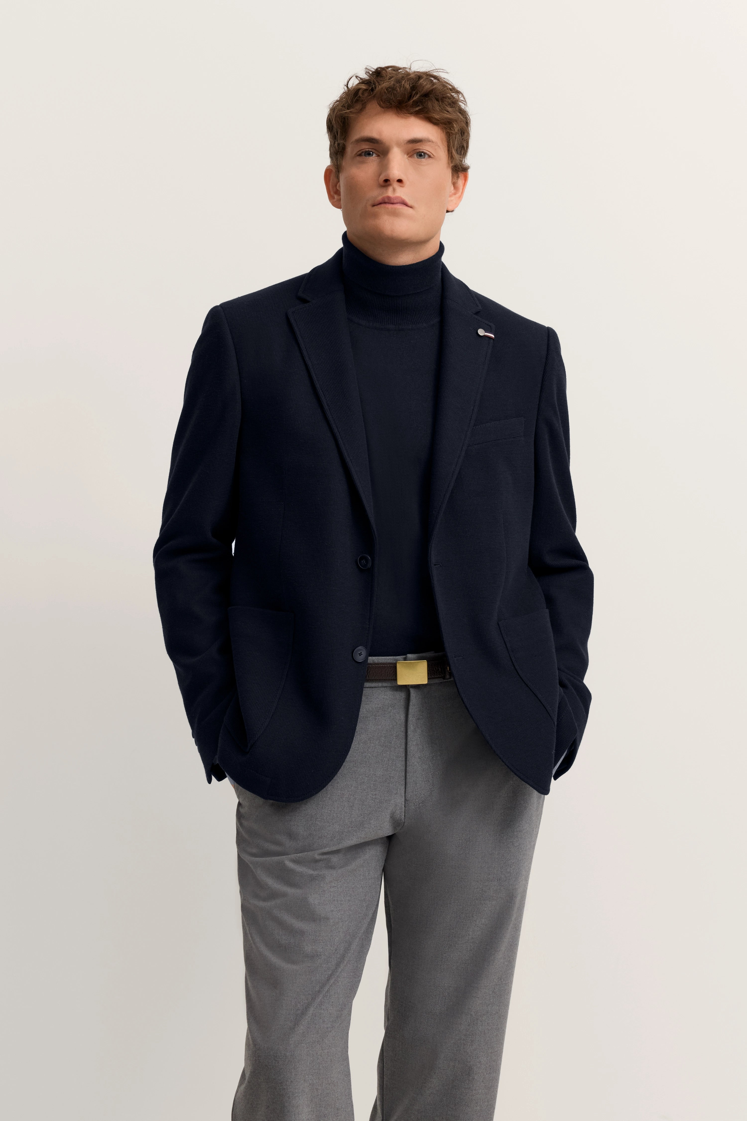 Model in navy bugatti Blazer, dunkelblauem Rollkragen und grauer Hose, Hände in Taschen, entspannte Haltung.