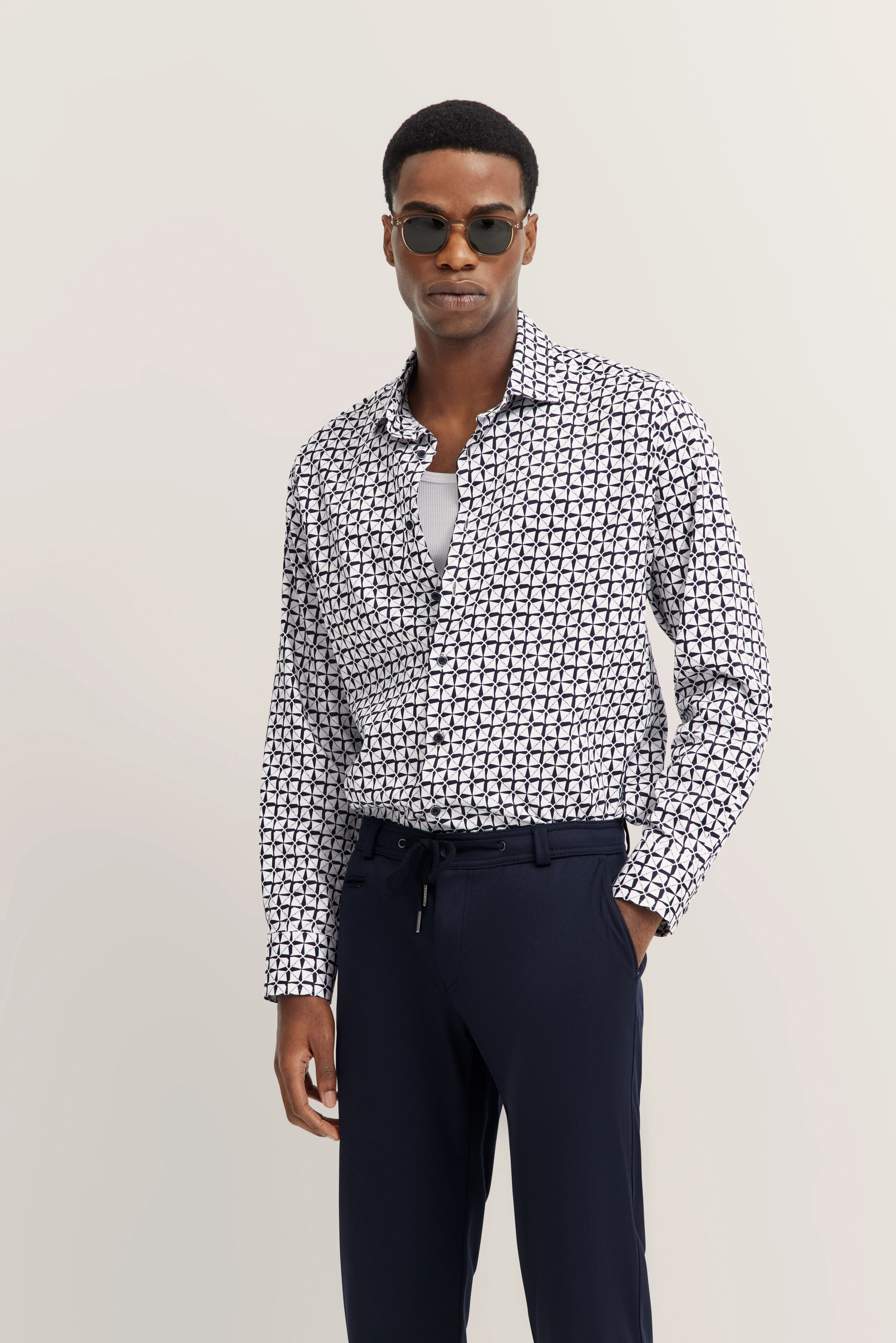 Man in een patroon overhemd en donkerblauwe broek, model staat ontspannen, draagt zonnebril.