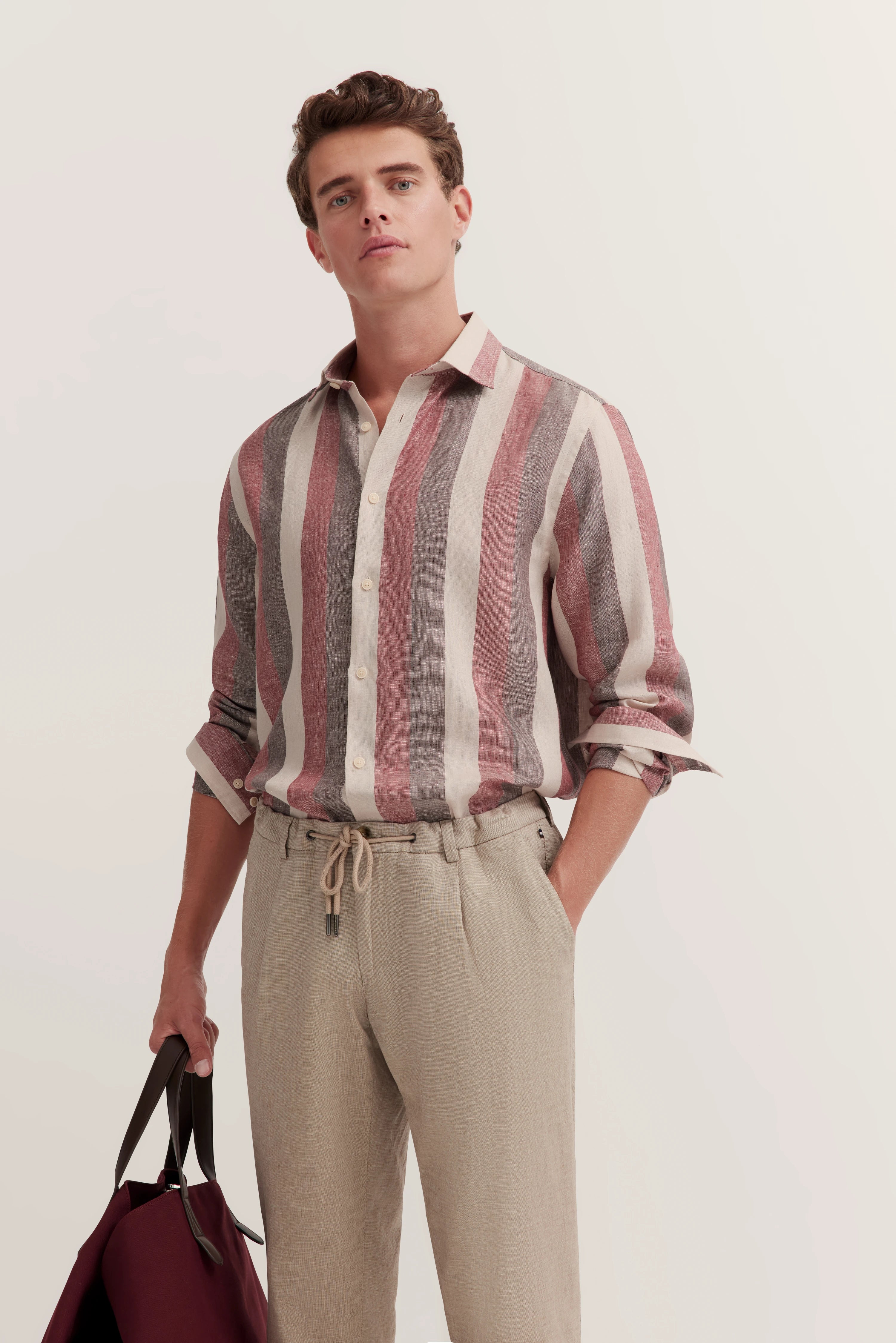 bugatti model in rood-beige gestreept shirt en beige broek, casual pose, tas in hand.