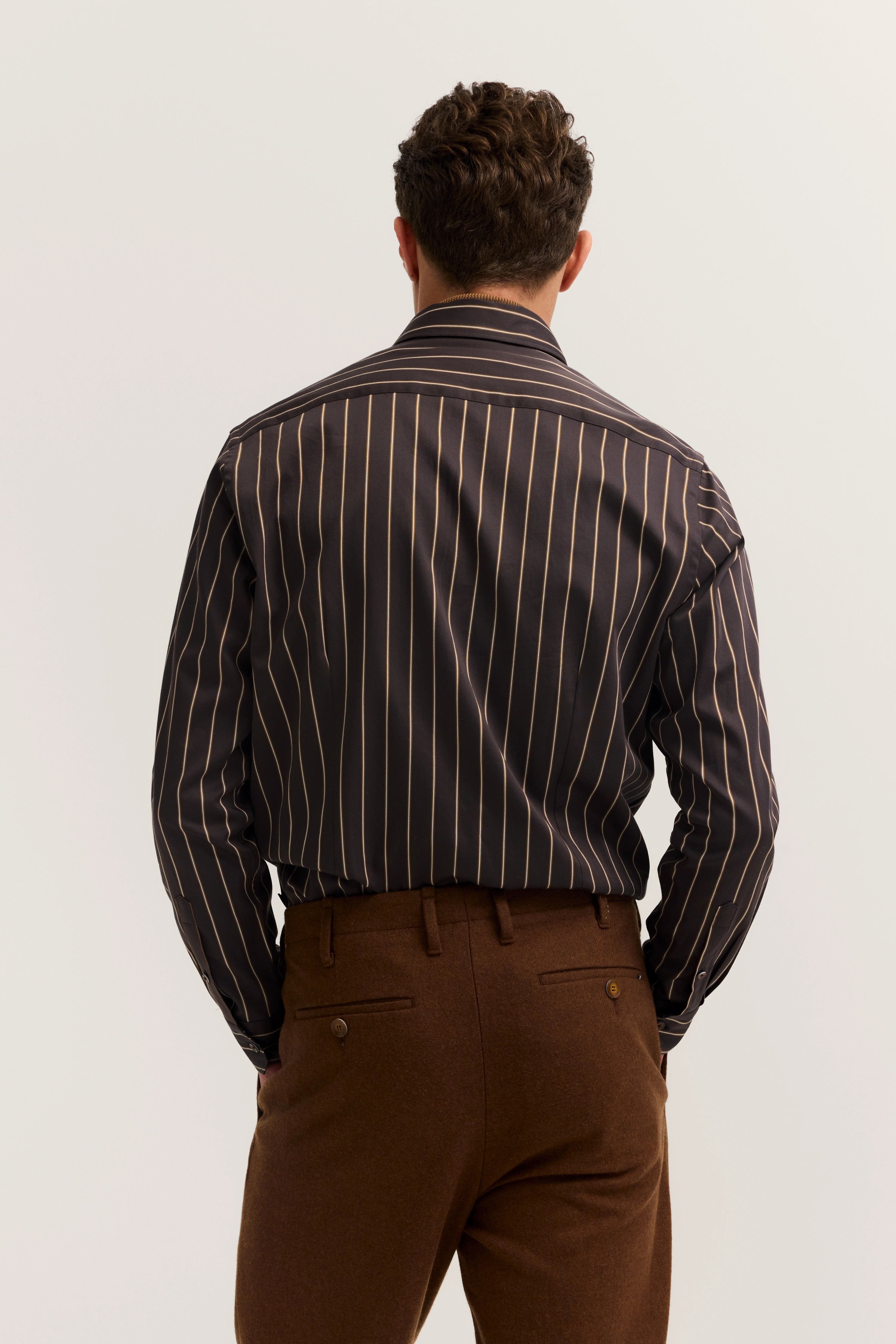 bugatti Hemd mit braunen Streifen, braune Hose. Rückansicht Model stehend, Hände in Hosentaschen.