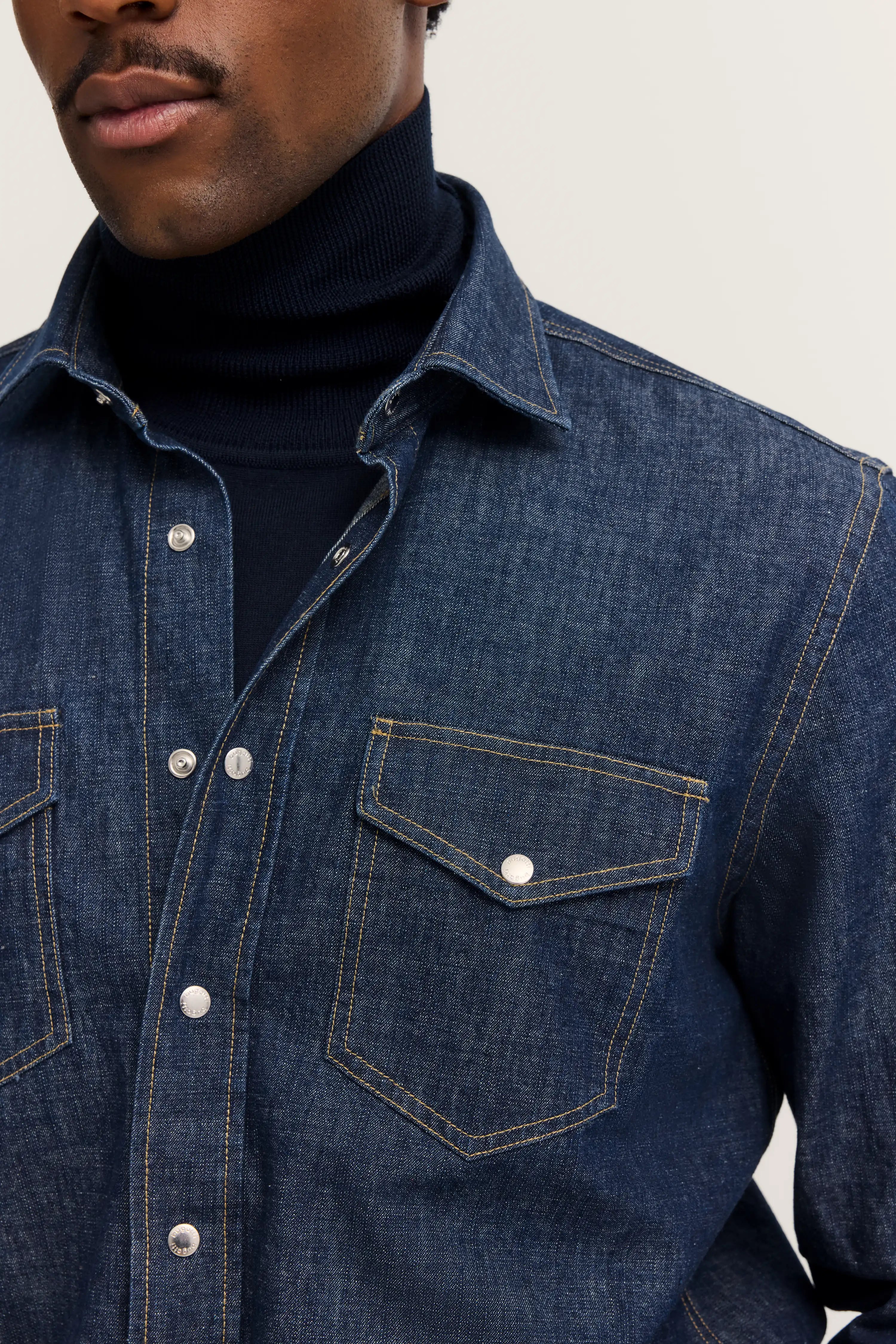 Denimhemd in Dunkelblau über schwarzem Rollkragenpullover, Büstenbild.