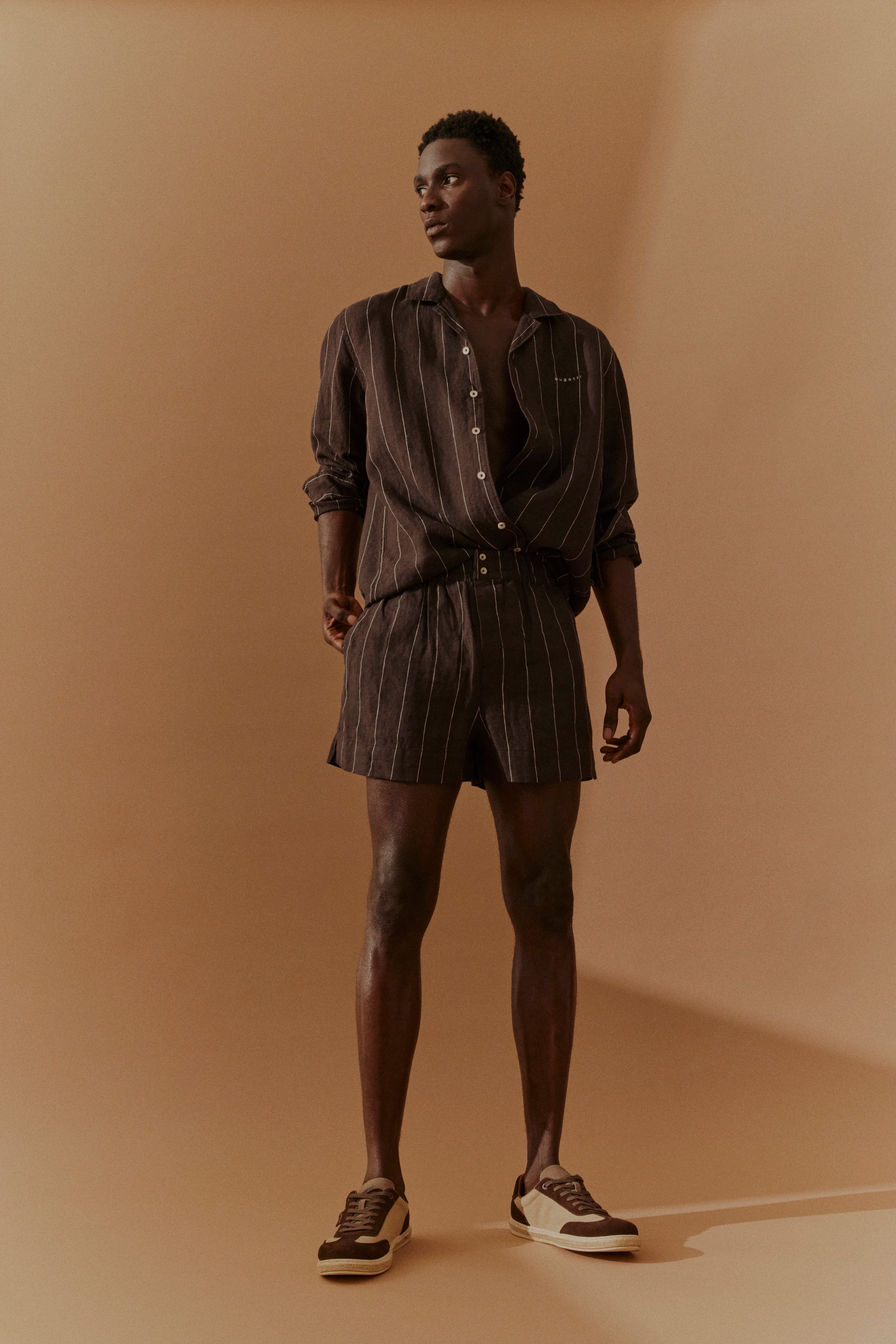 Modelbild in braunem Nadelstreifen-Zweiteiler mit offenem Hemd, lässige Pose, bugatti Stil.