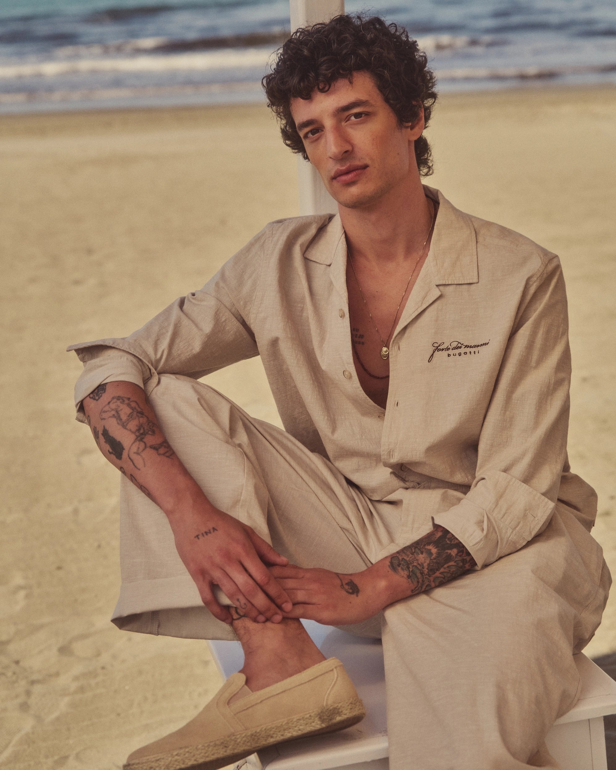Beige bugatti Leinenoutfit, Model am Strand, sitzt entspannt, sommerlicher Look, ruht lässig auf Stuhl.