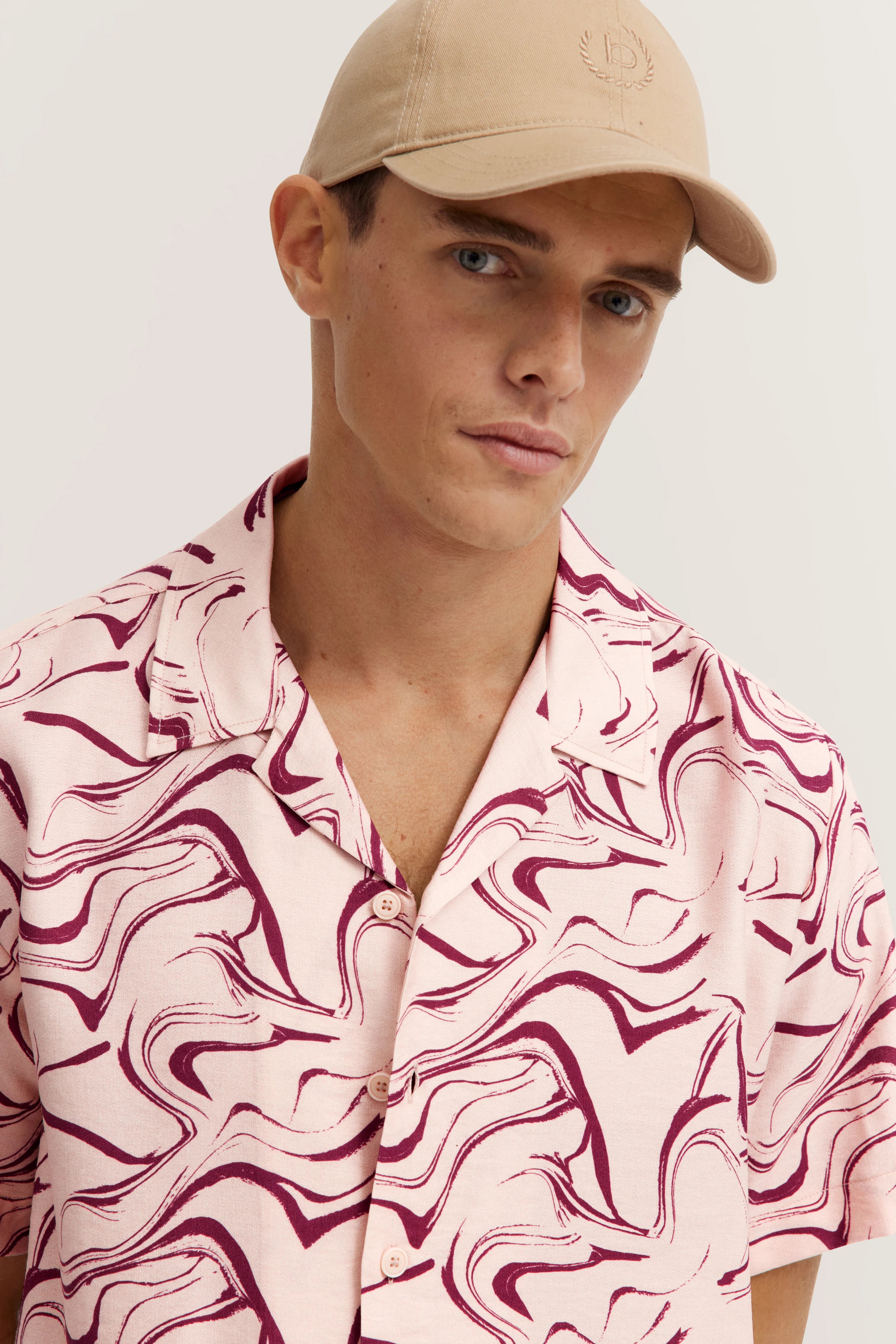 Model in roze gemonteerd shirt en beige pet, ontspannen houding.
