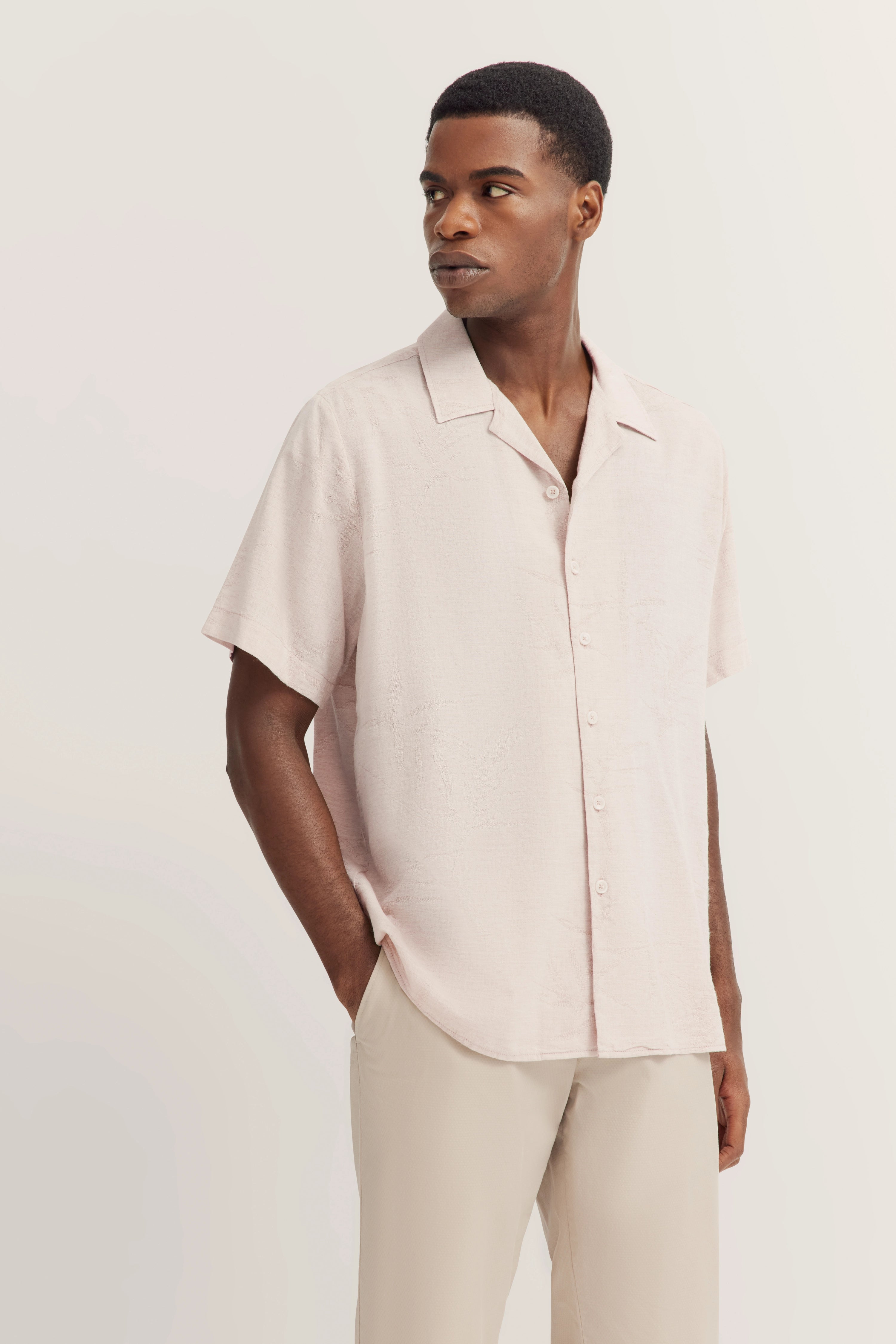 Model in beige, kort mouw shirt en bijpassende broek. Ontspannen pose, handen in de zakken.