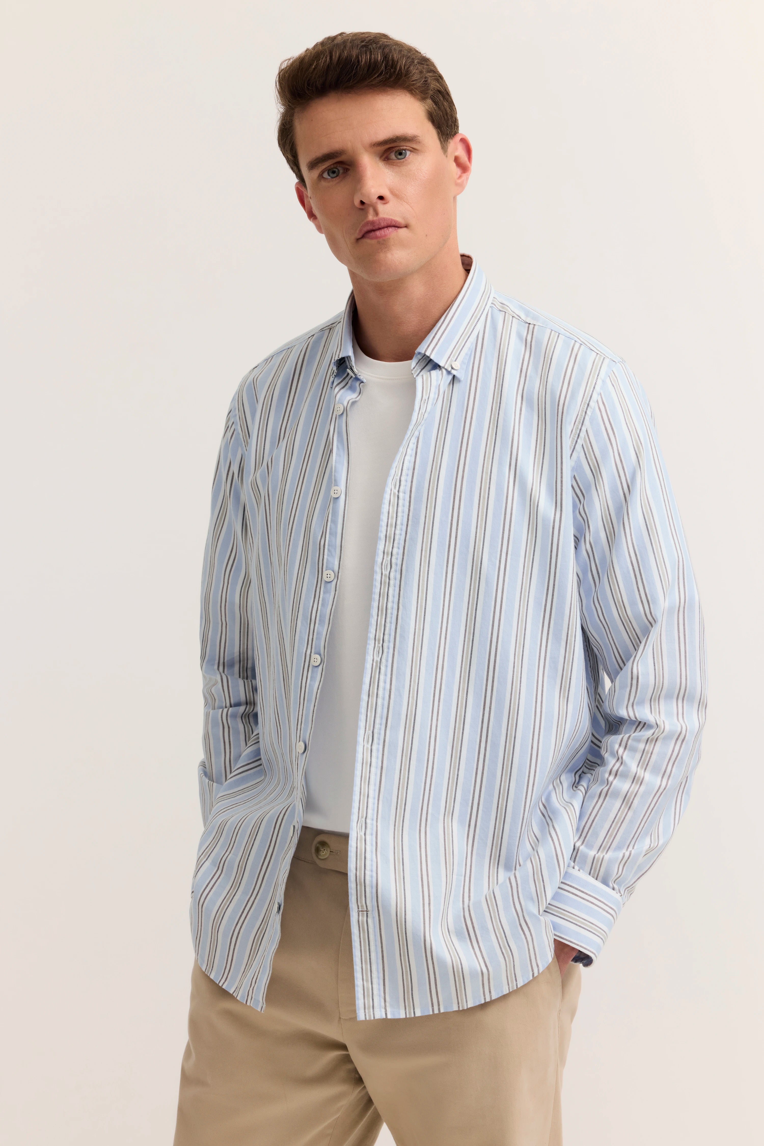 Blau-weiß gestreiftes Hemd über weißem Shirt, beige Hose, legeres Modelbild, Hände in Hosentaschen.