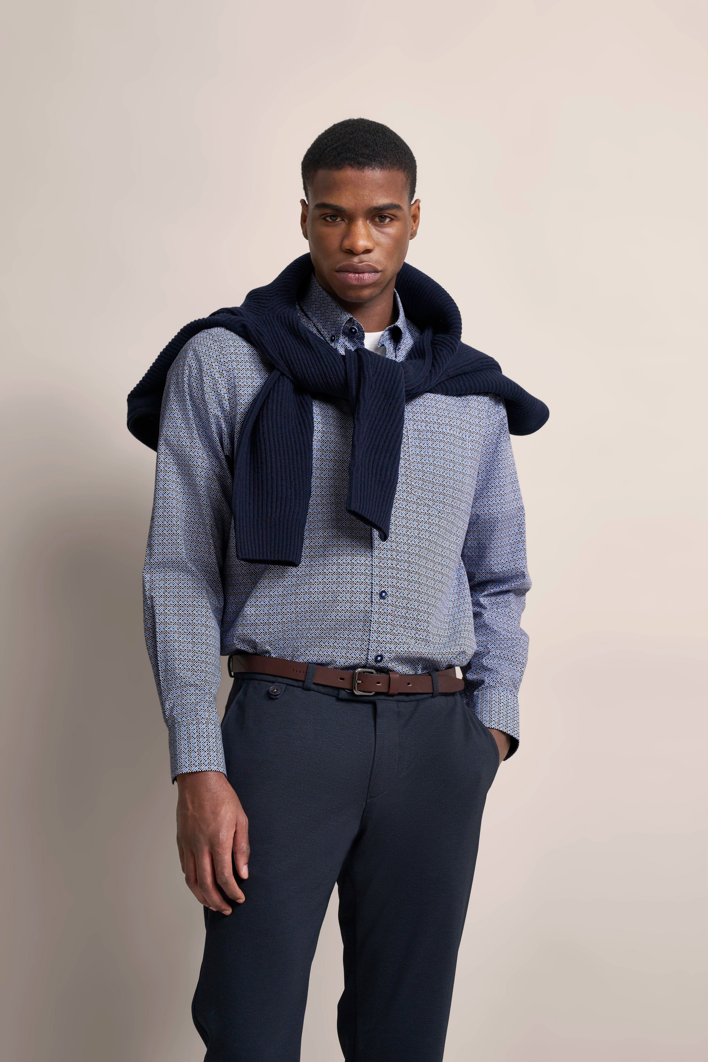 Model in gemustertem Hemd, dunkelblauer Pullover über Schultern, blaue Hose