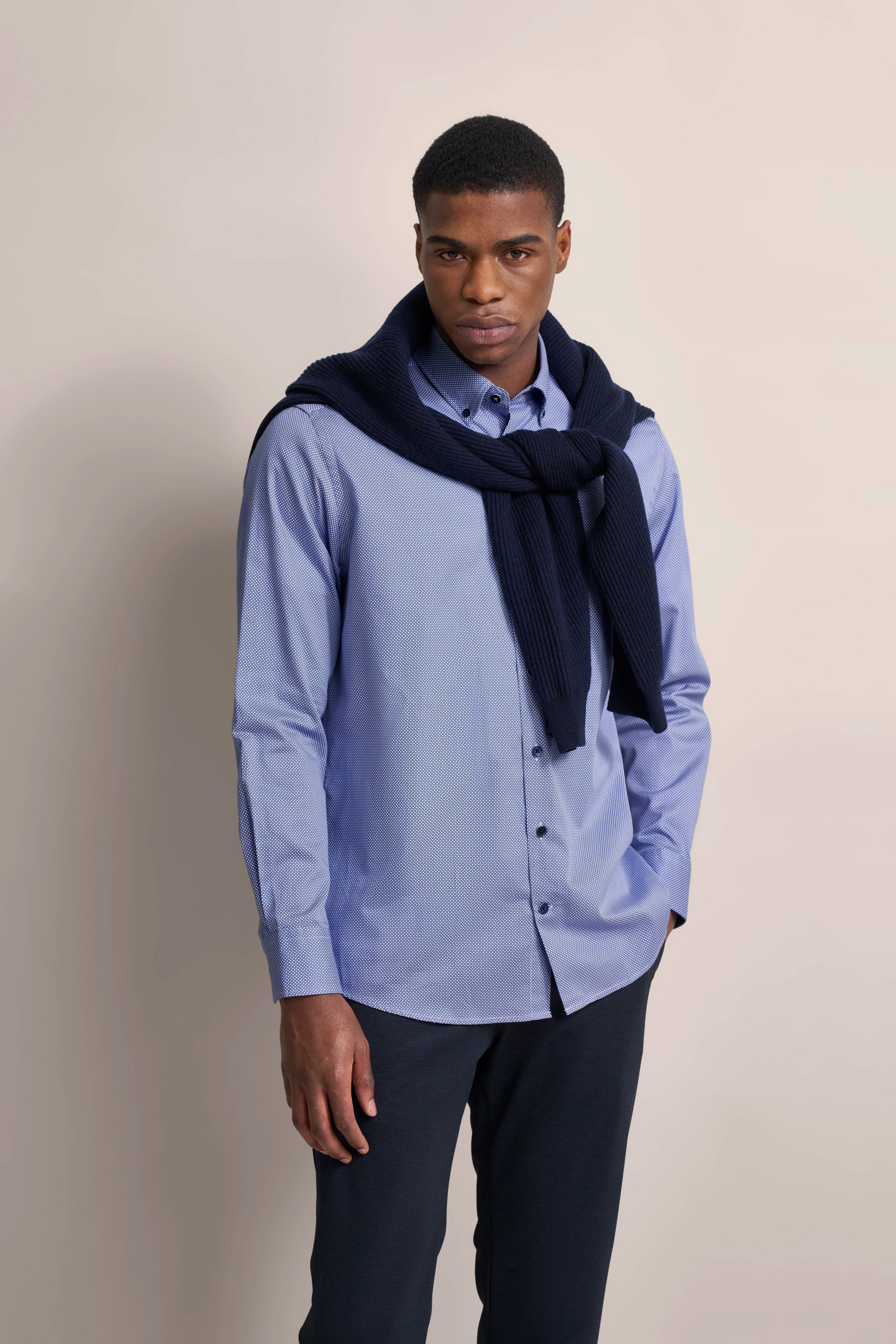 Model trägt blaues Hemd mit Muster, dunkelblauer Pullover um die Schultern, schwarze Hose.