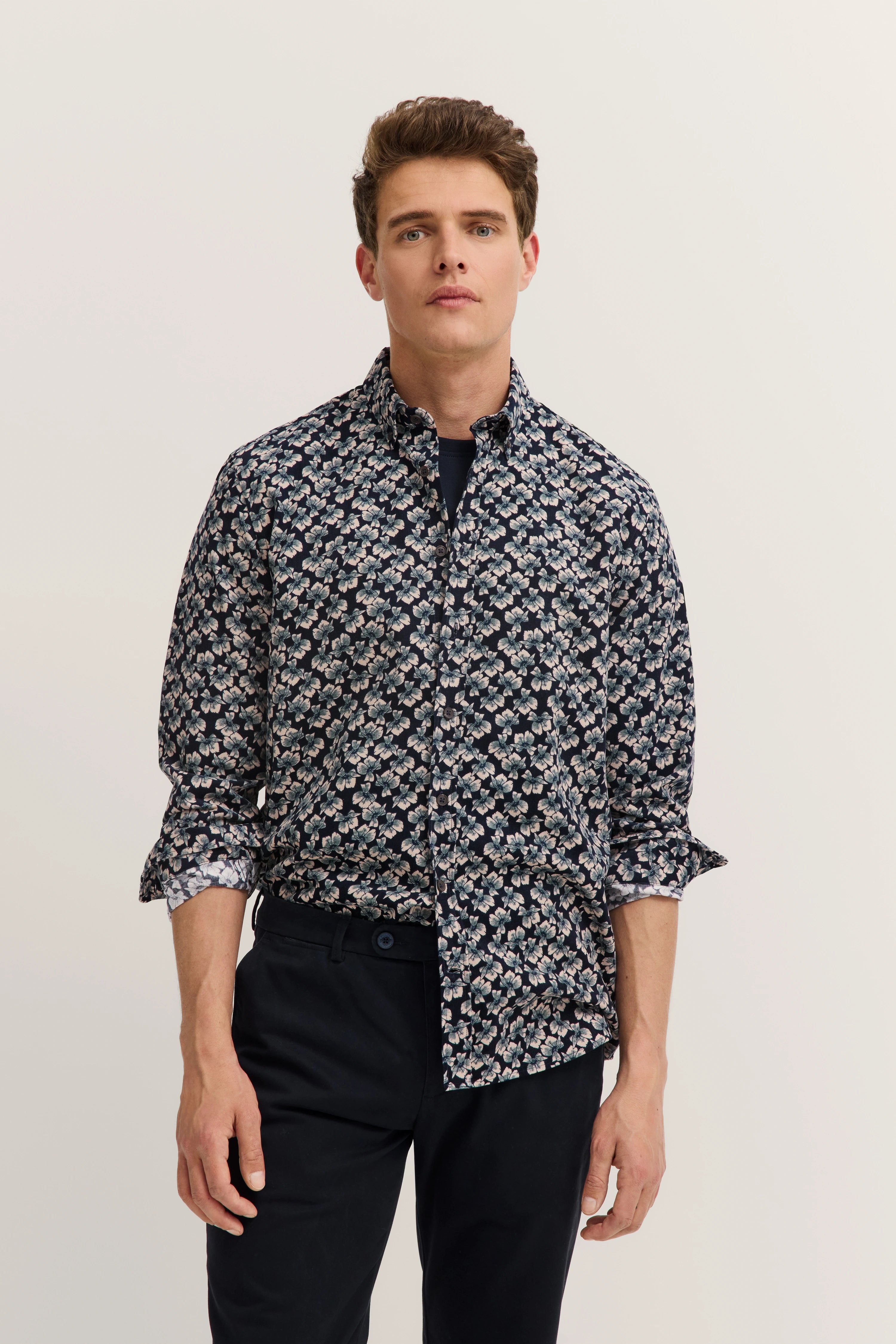 Model in bloemig bugatti overhemd, donkere achtergrond, ontspannen houding, zwarte broek, elegantie en stijl.