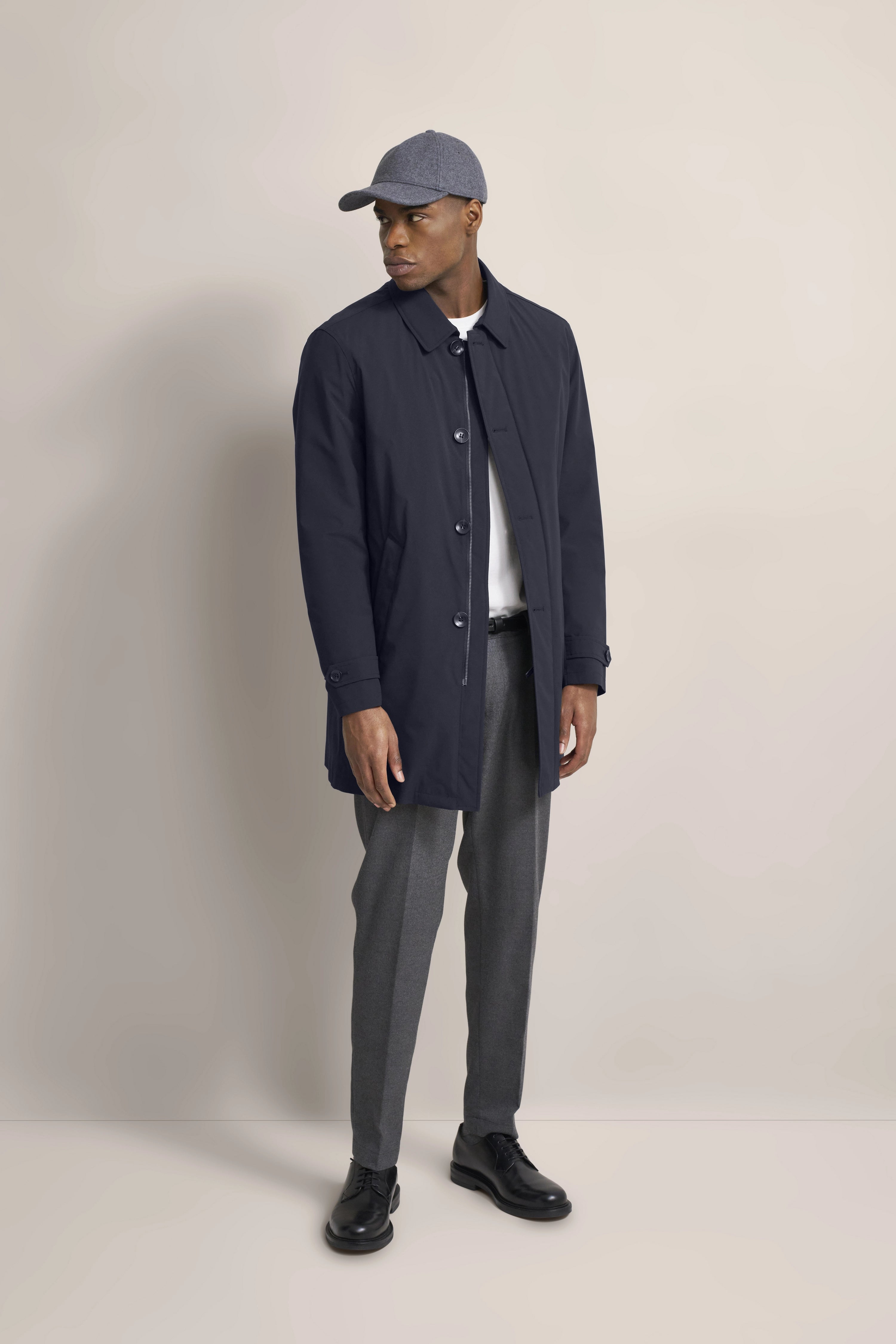 Model in navy bugatti coat, gray pants, and cap, casual pose.