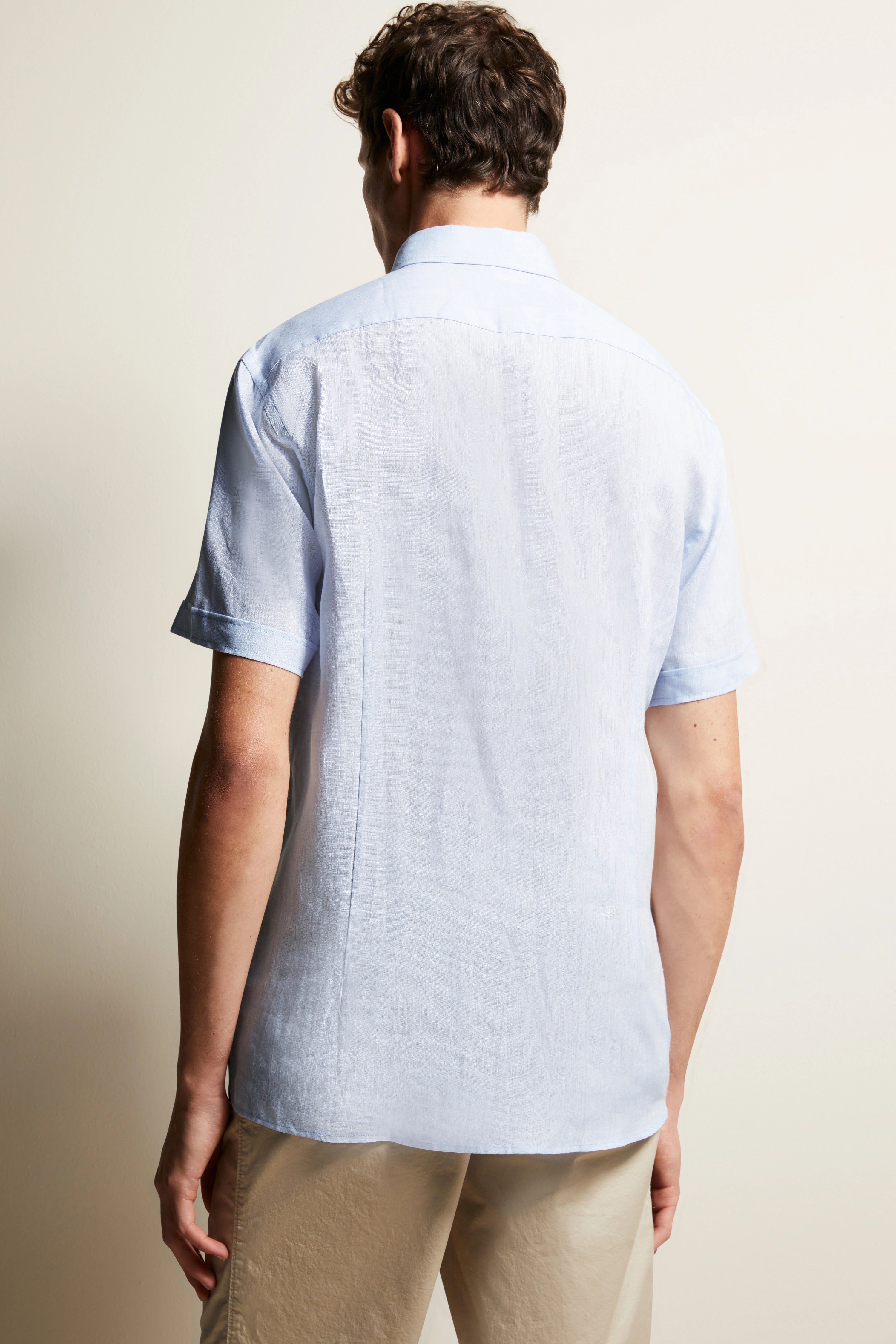 Achteraanzicht model met lichtblauw bugatti overhemd met korte mouwen, beige broek, casual pose.