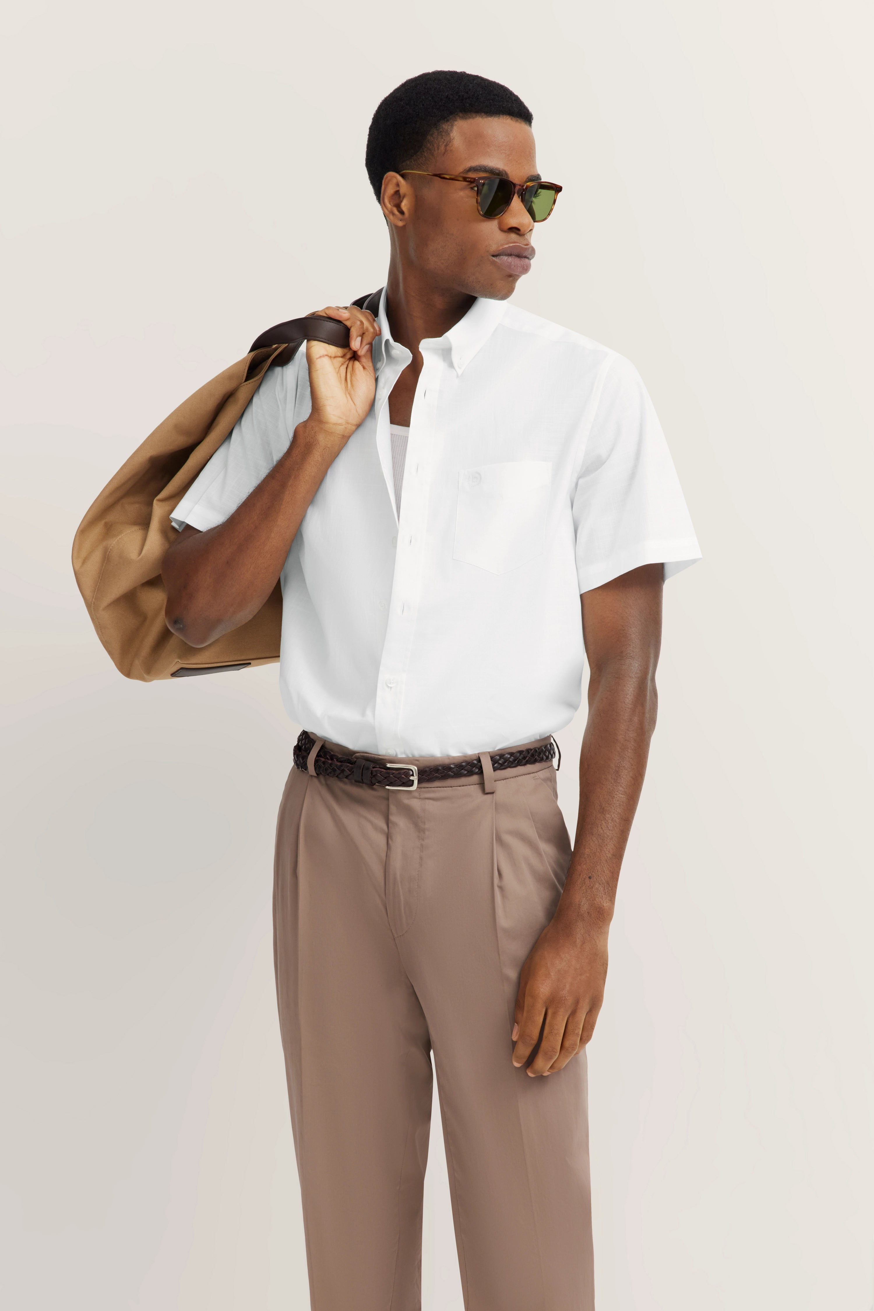 Model in wit overhemd, beige broek, houdt jas over schouder, zonnebril, casual pose.