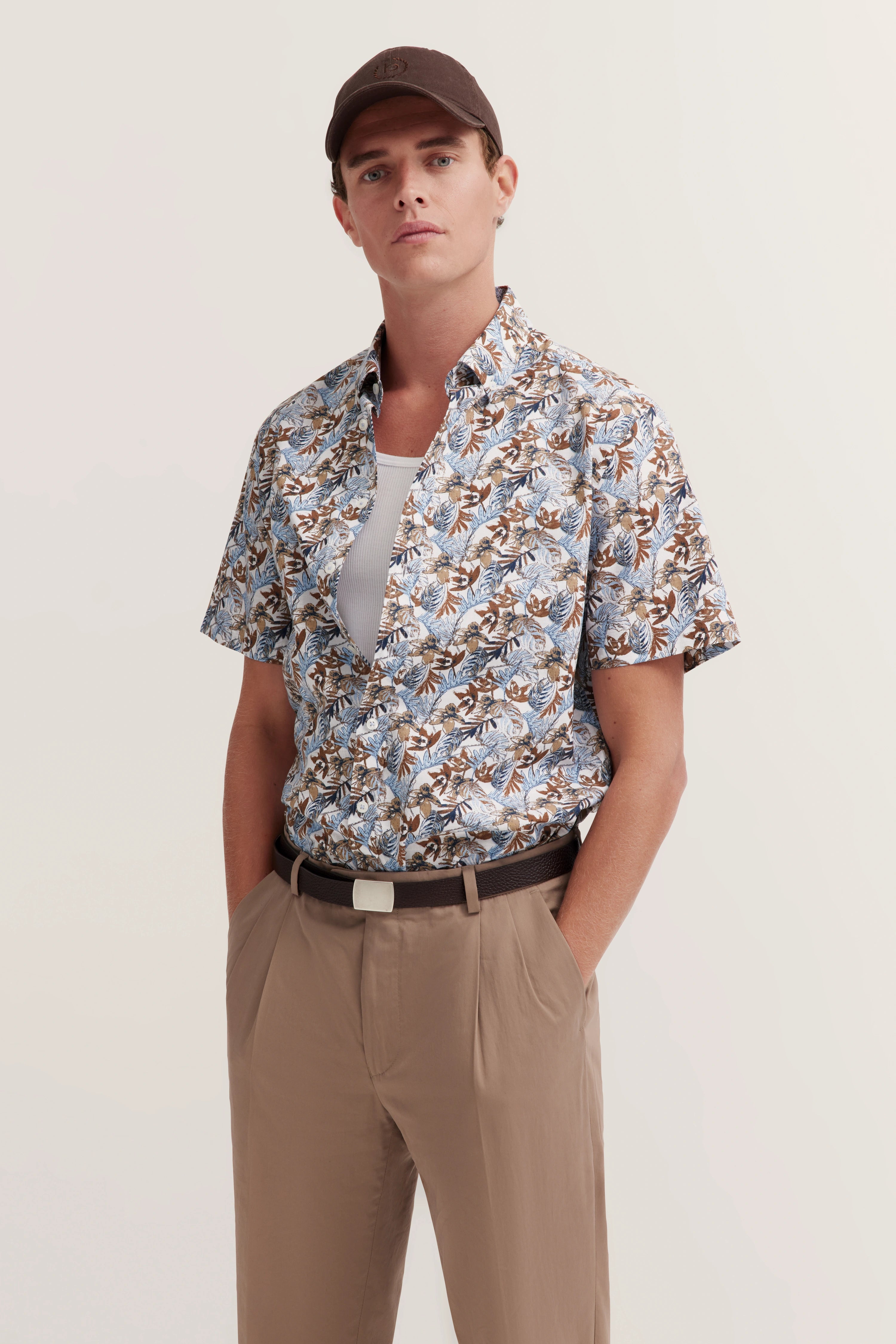Model in bloemrijk bugatti overhemd, beige broek, bruine pet, ontspannen houding, handen in zakken.