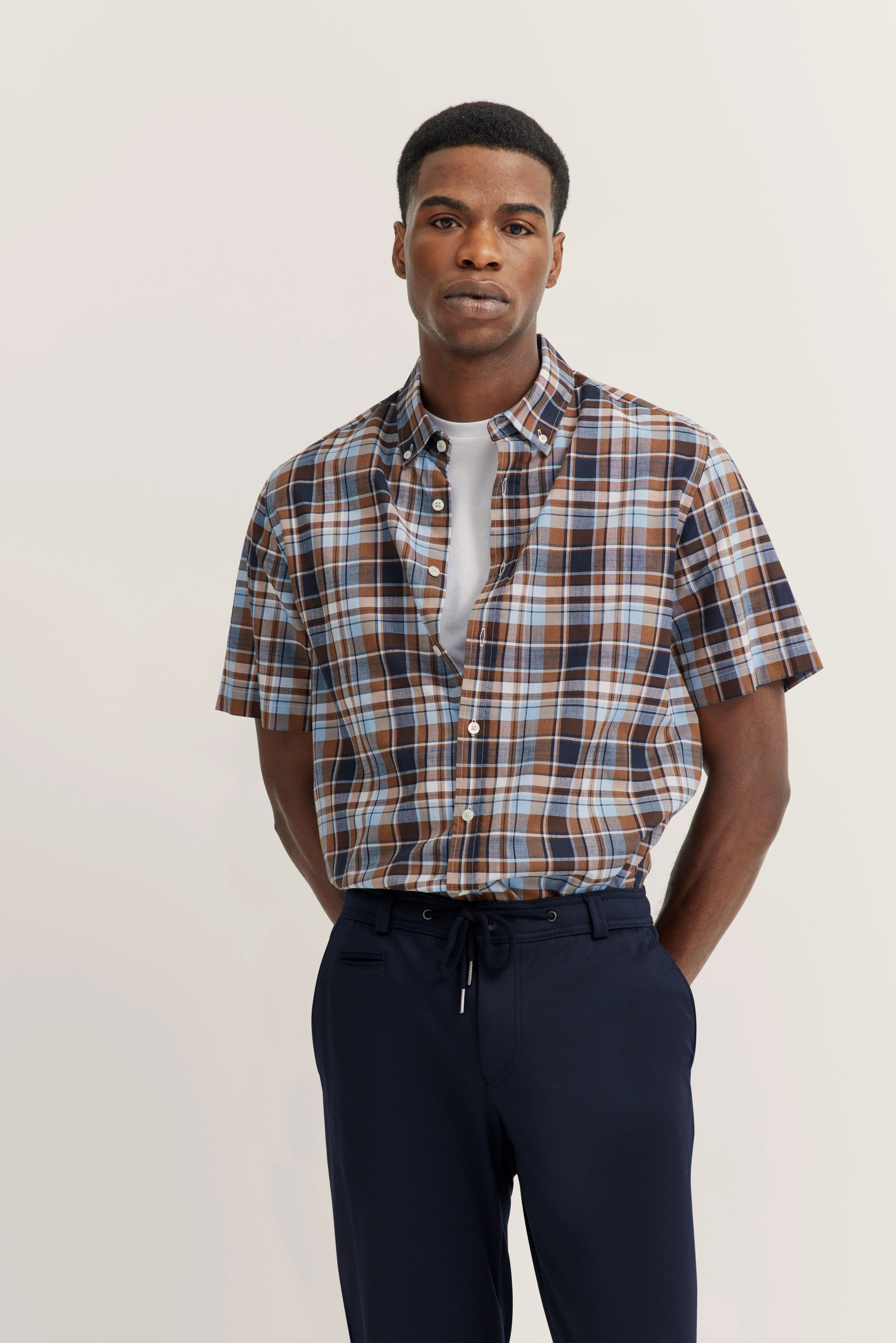 Geruit kort mouw shirt in blauw-bruin over wit shirt, donkere broek, model staat ontspannen.