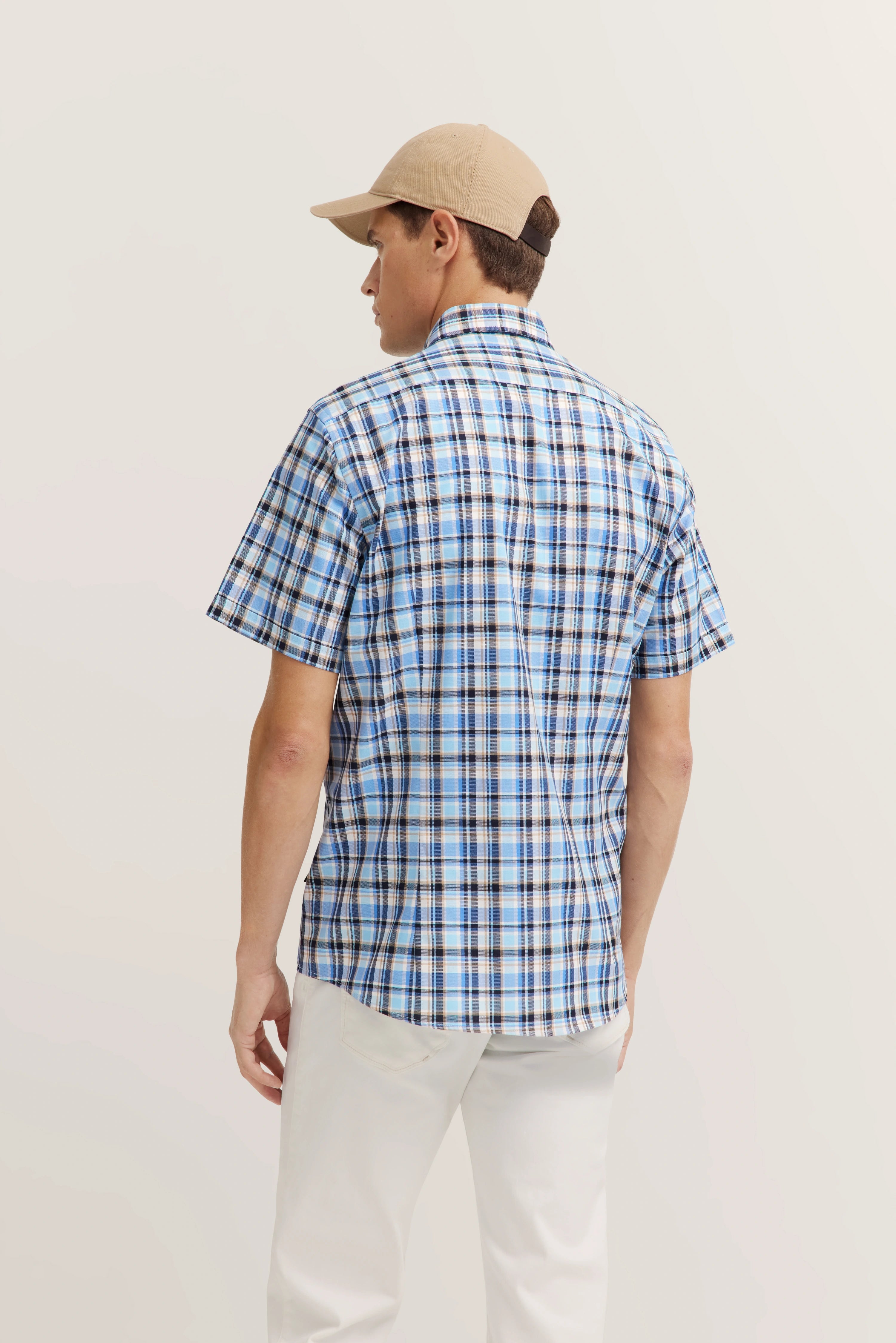 Man in blauw geruit bugatti-overhemd, ontspannen stijl, achterkant, beige pet, witte broek.