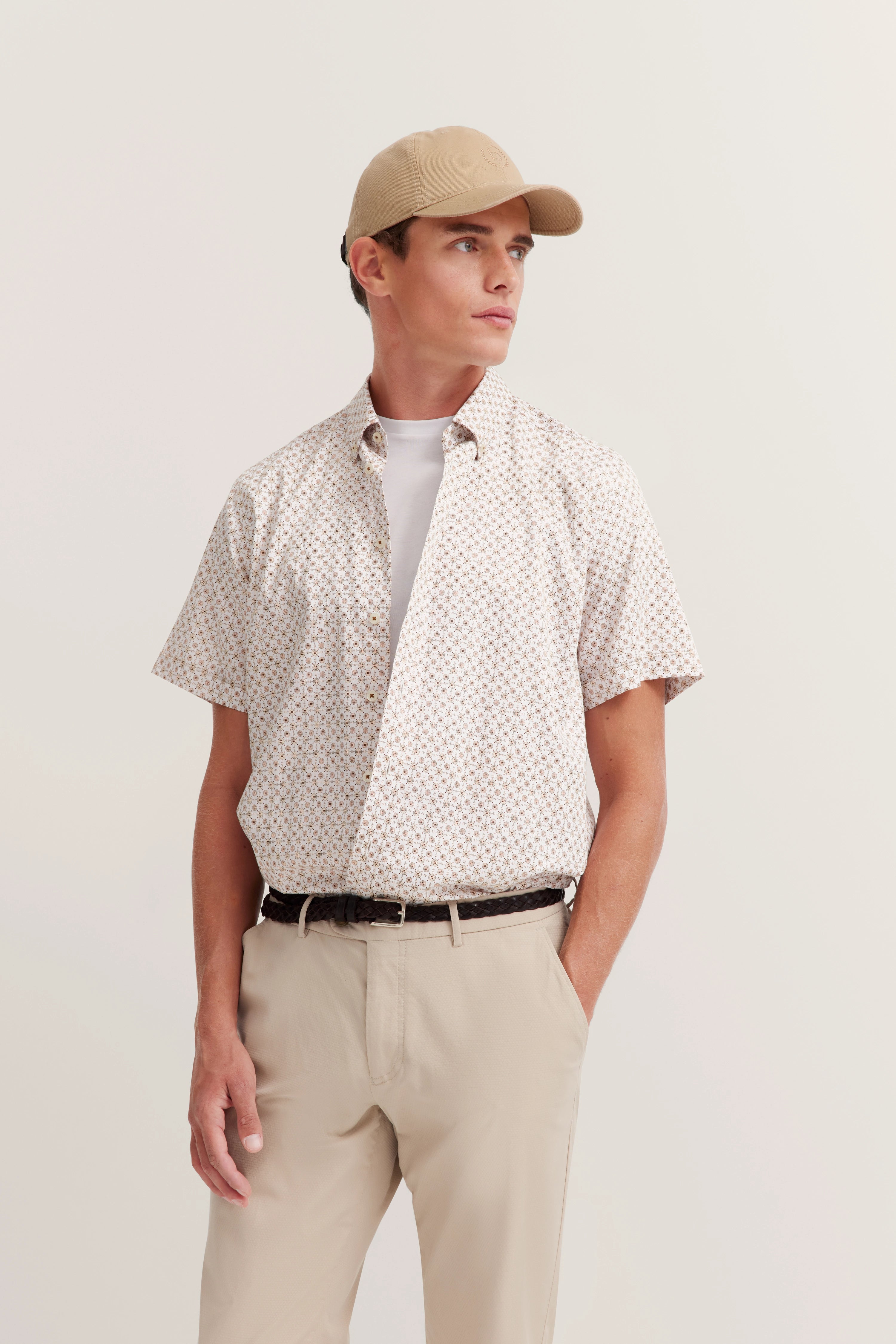 Model in beige outfit met een bedrukt kortemouwenshirt, wit T-shirt, beige broek en pet.
