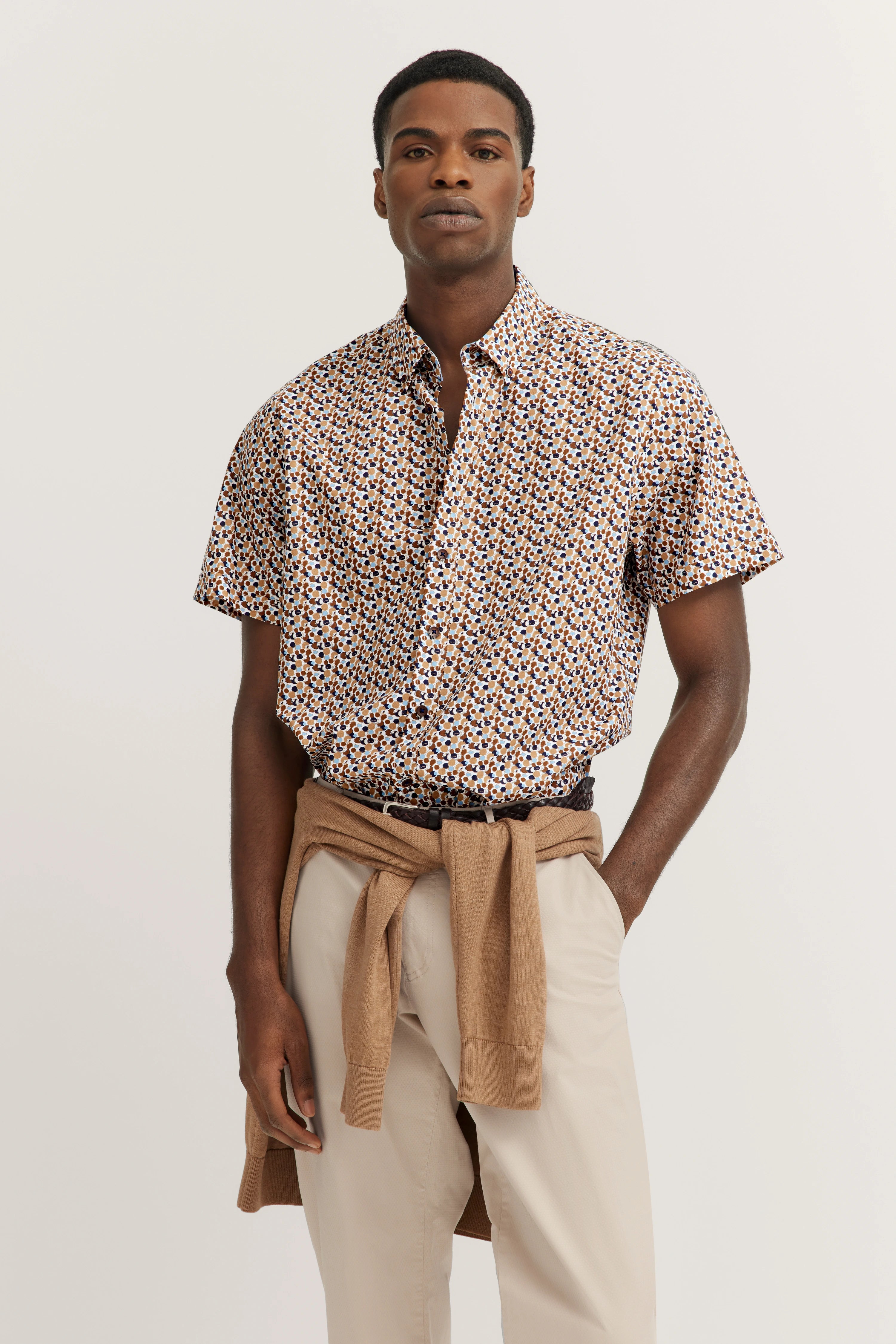 Model in kortem overhemd met patroon, beige broek, trui om de heupen, ontspannen houding.