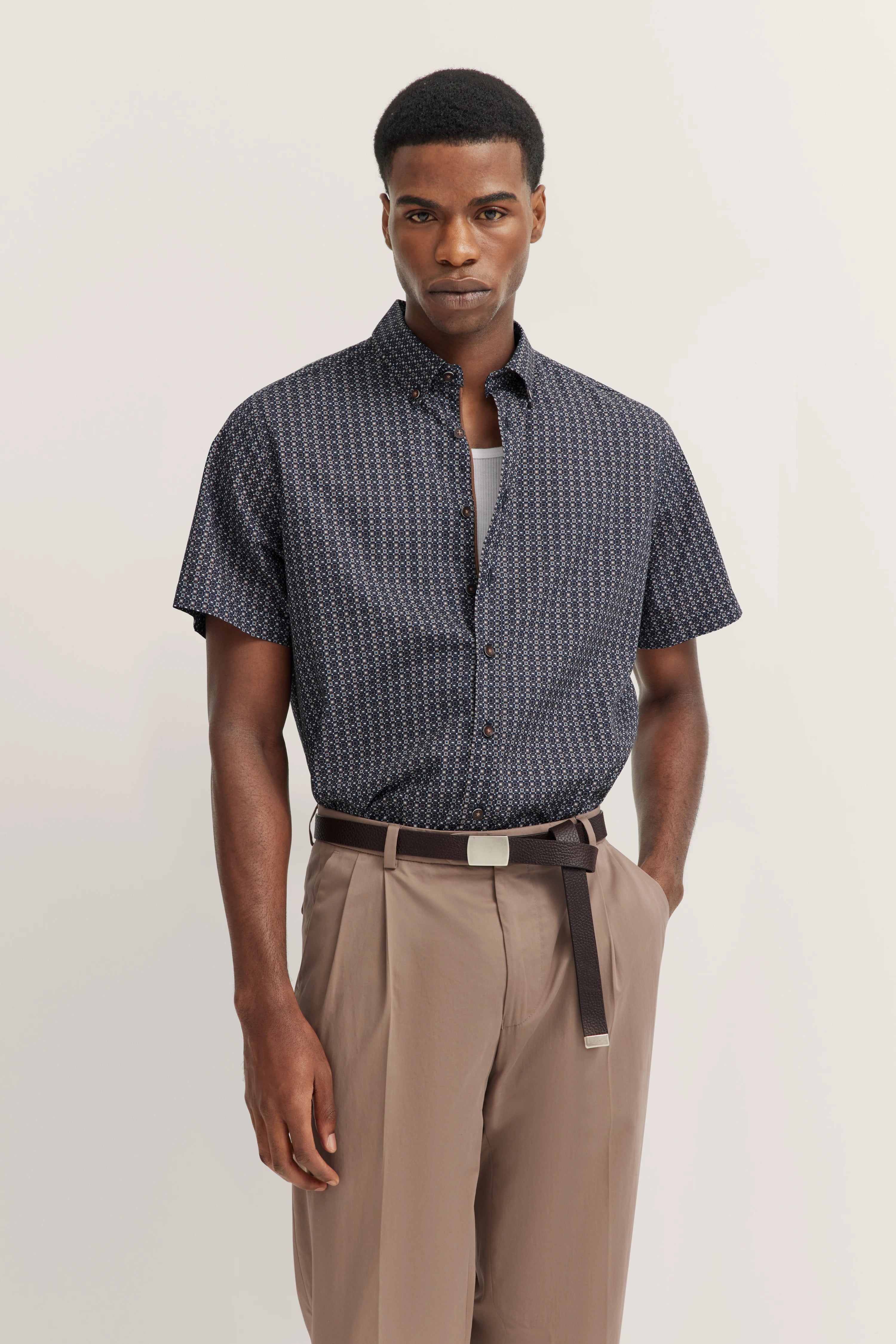 Donker gemonteerd overhemd, beige broek, model staand, handen in zakken.