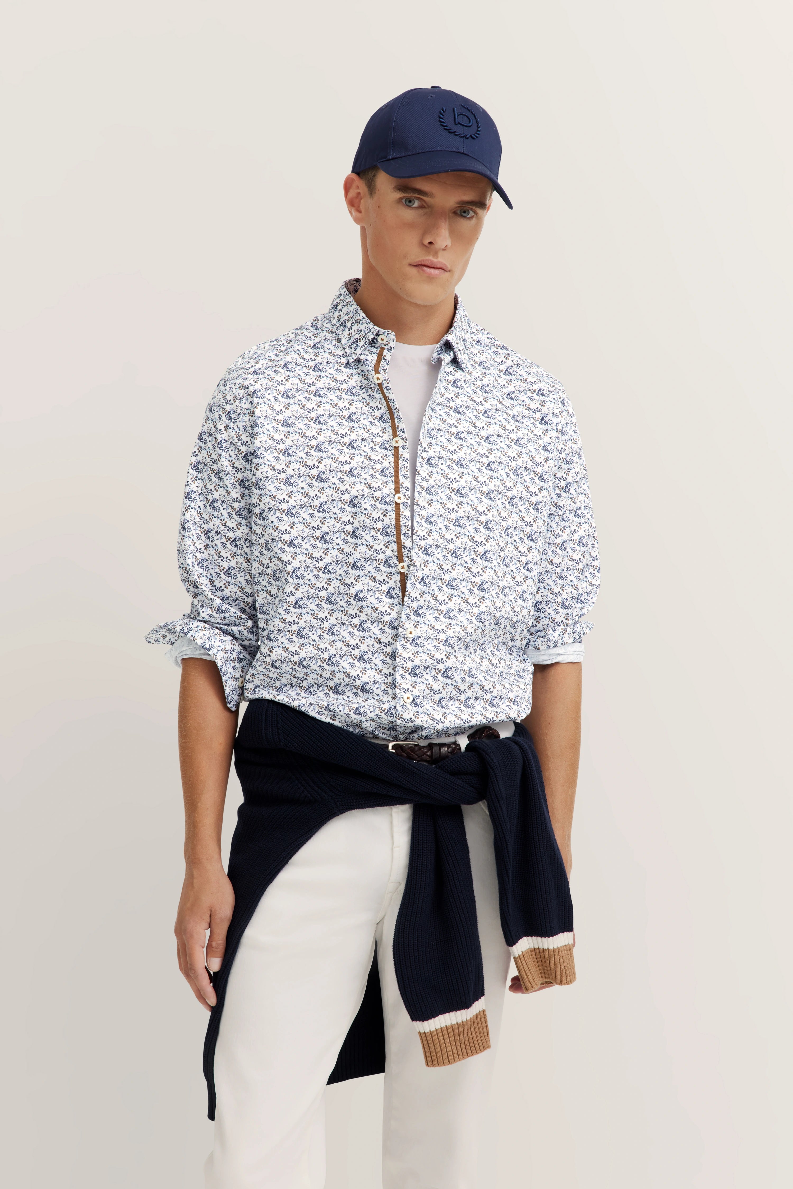 Model in een gedessineerde blouse, wit shirt, beige riem, witte broek, donkerblauwe pet, ontspannen pose.