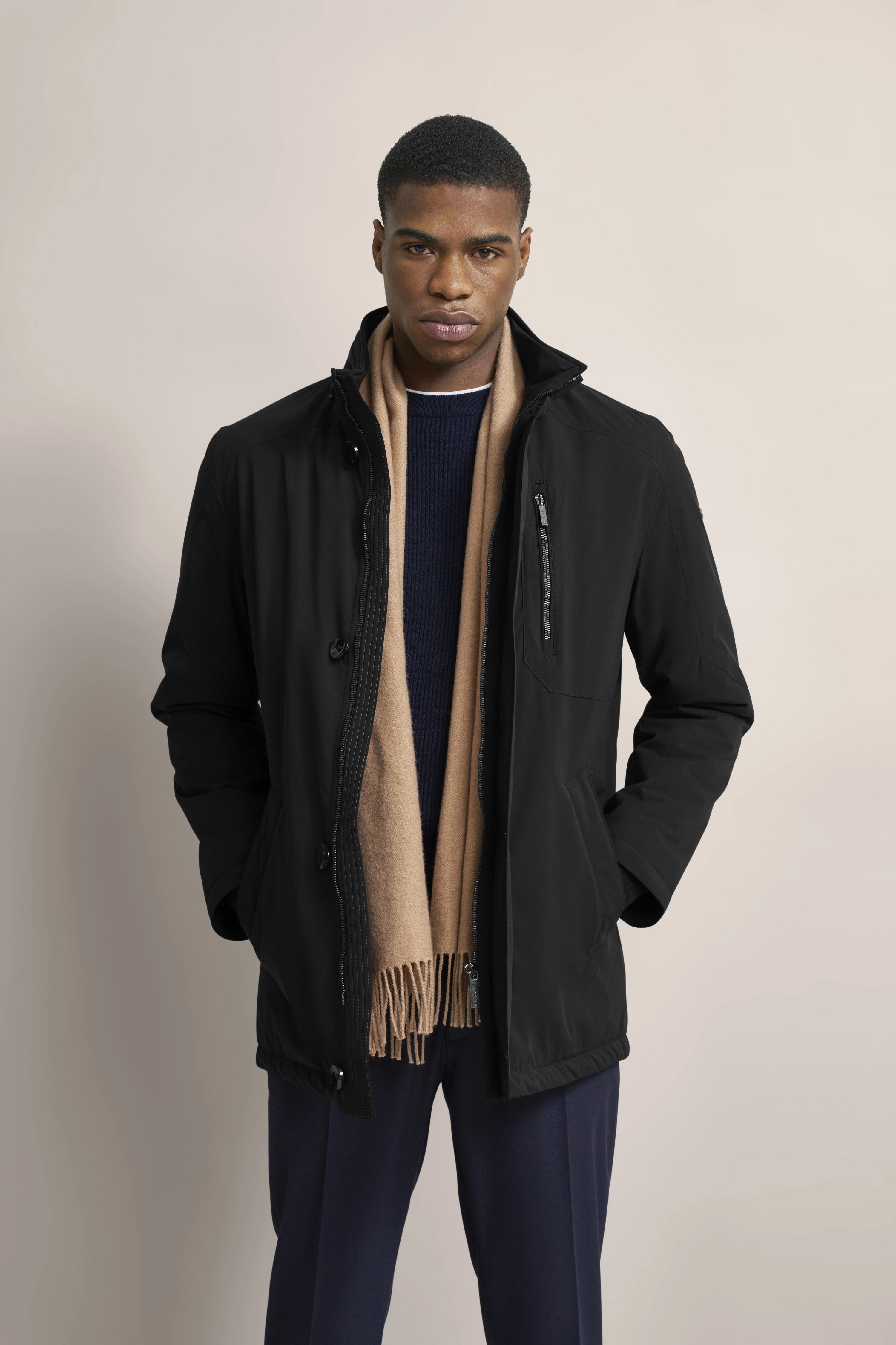 Modelbild: Schwarze Jacke, marineblauer Pullover, beige Schal. Stehendes Posing, Hände in den Taschen.