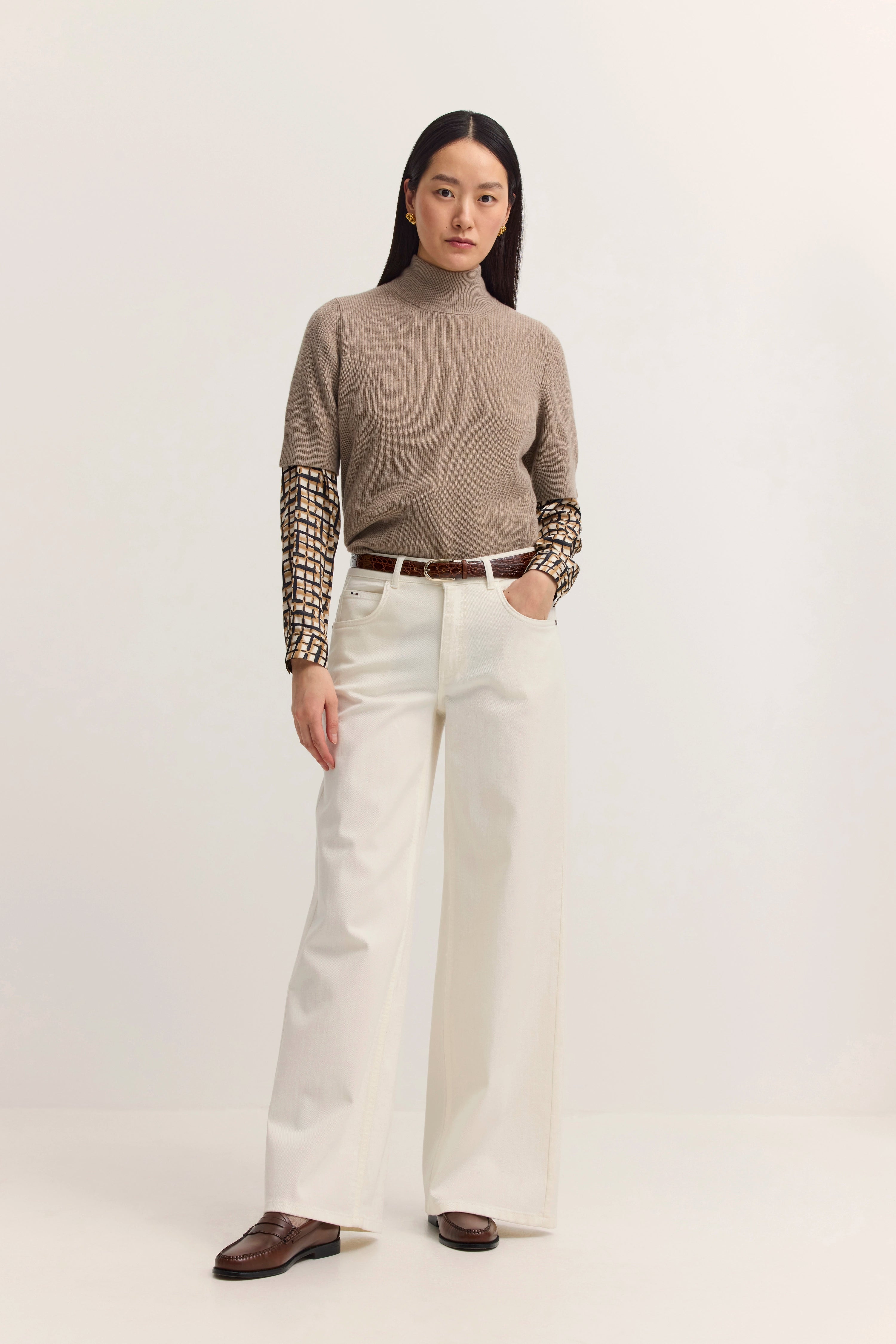 Model in beige Rollkragenpullover, weißer Hose und braunen Schuhen, stehende Pose.