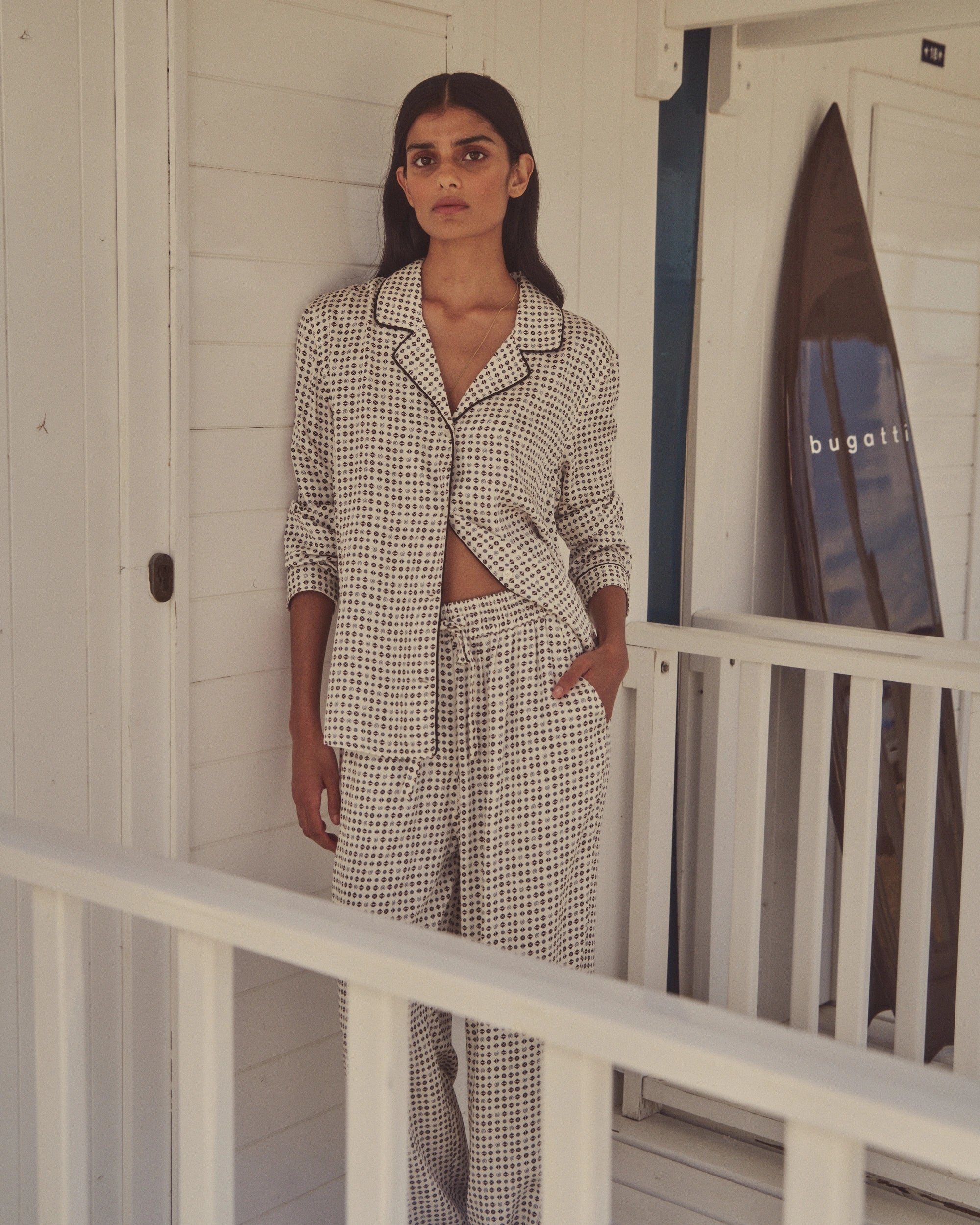 Model in gemustertem Pyjama-Set in Weiß/Schwarz, legeres Posing, bugatti Surfbrett im Hintergrund.