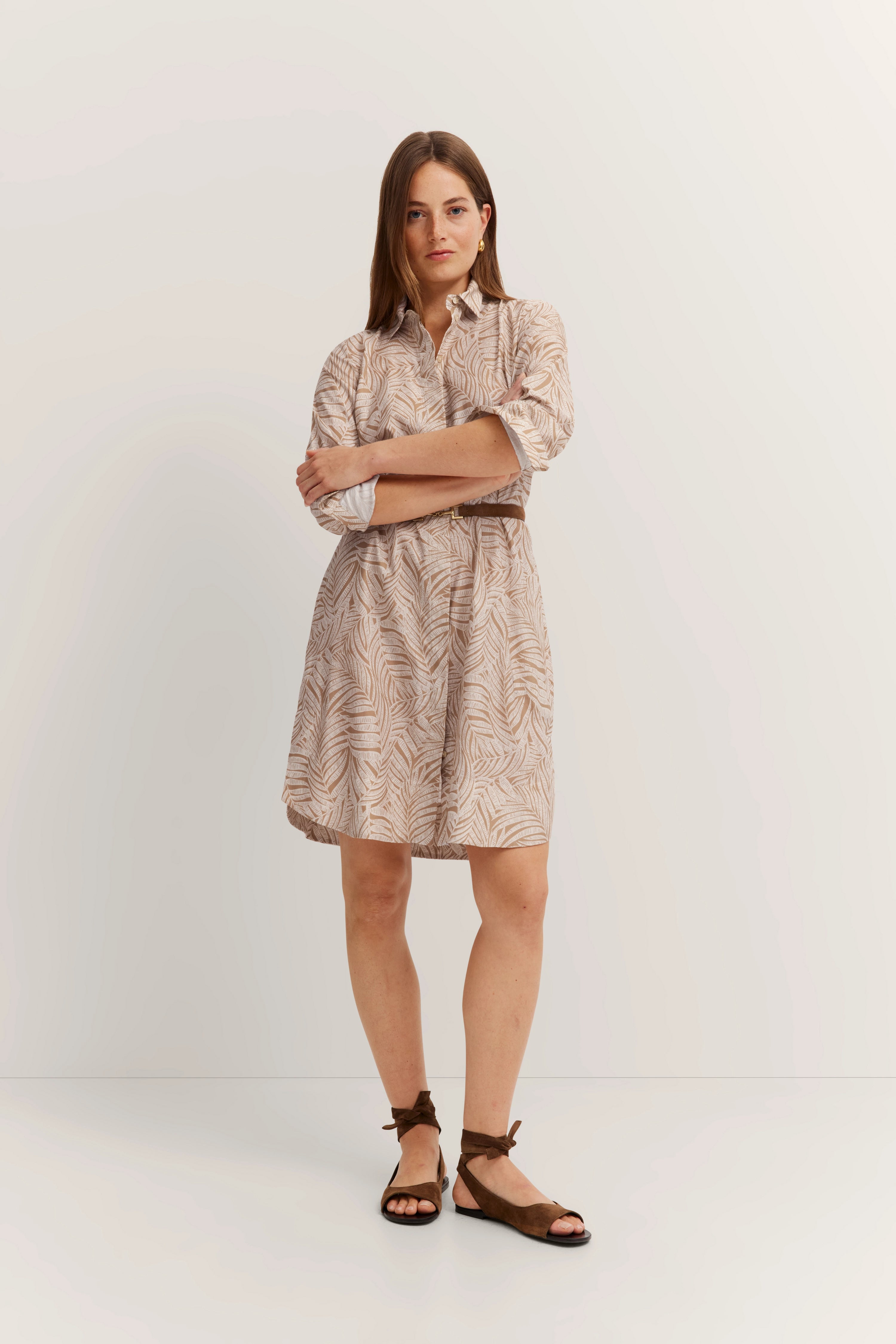 Model in beige, bedrukt jurkje met lange mouwen, armen gekruist, bruine sandalen.