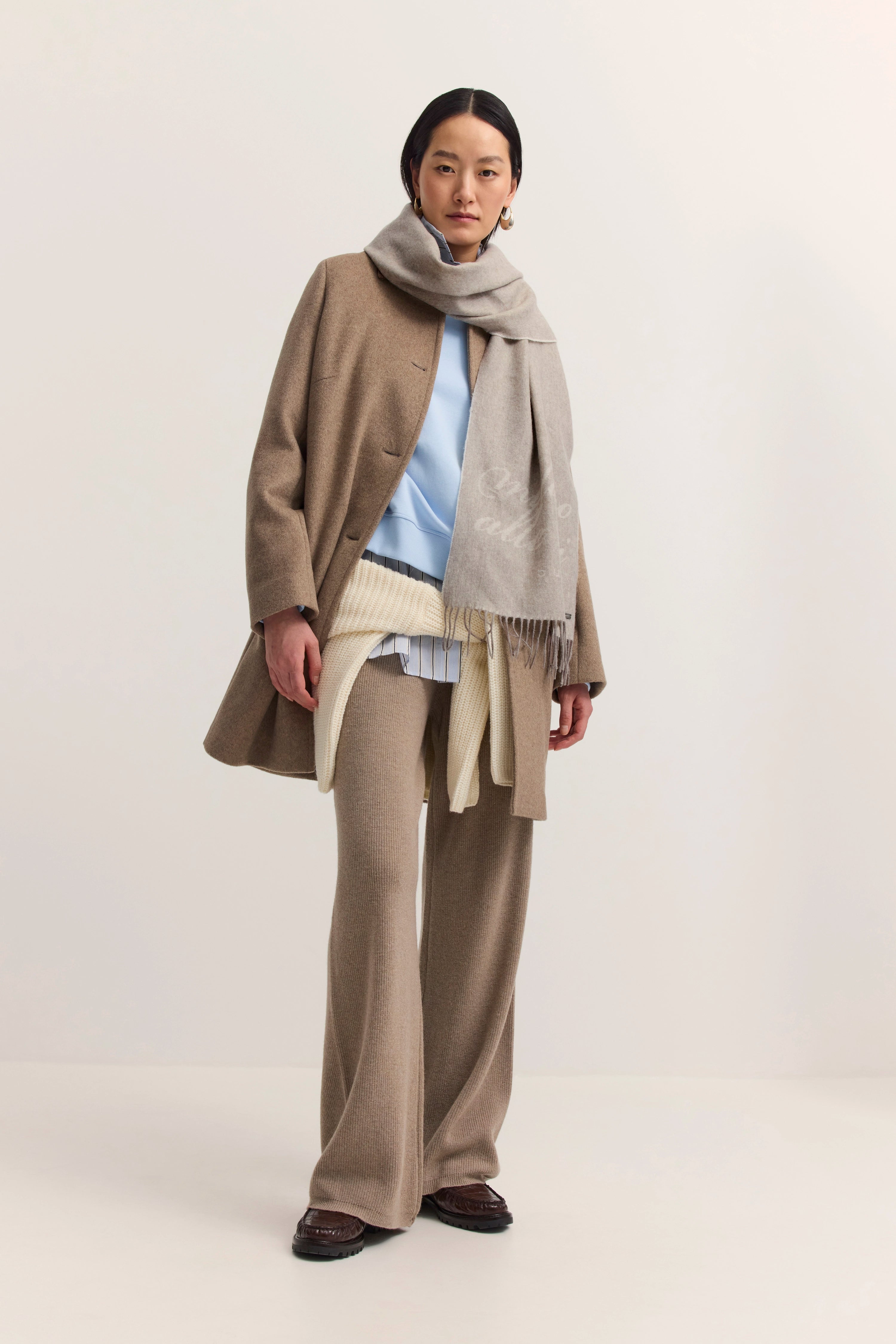 Elegantes Model mit beigem Mantel, grauem Schal, blauem Pulli, beigefarbener Hose von bugatti.