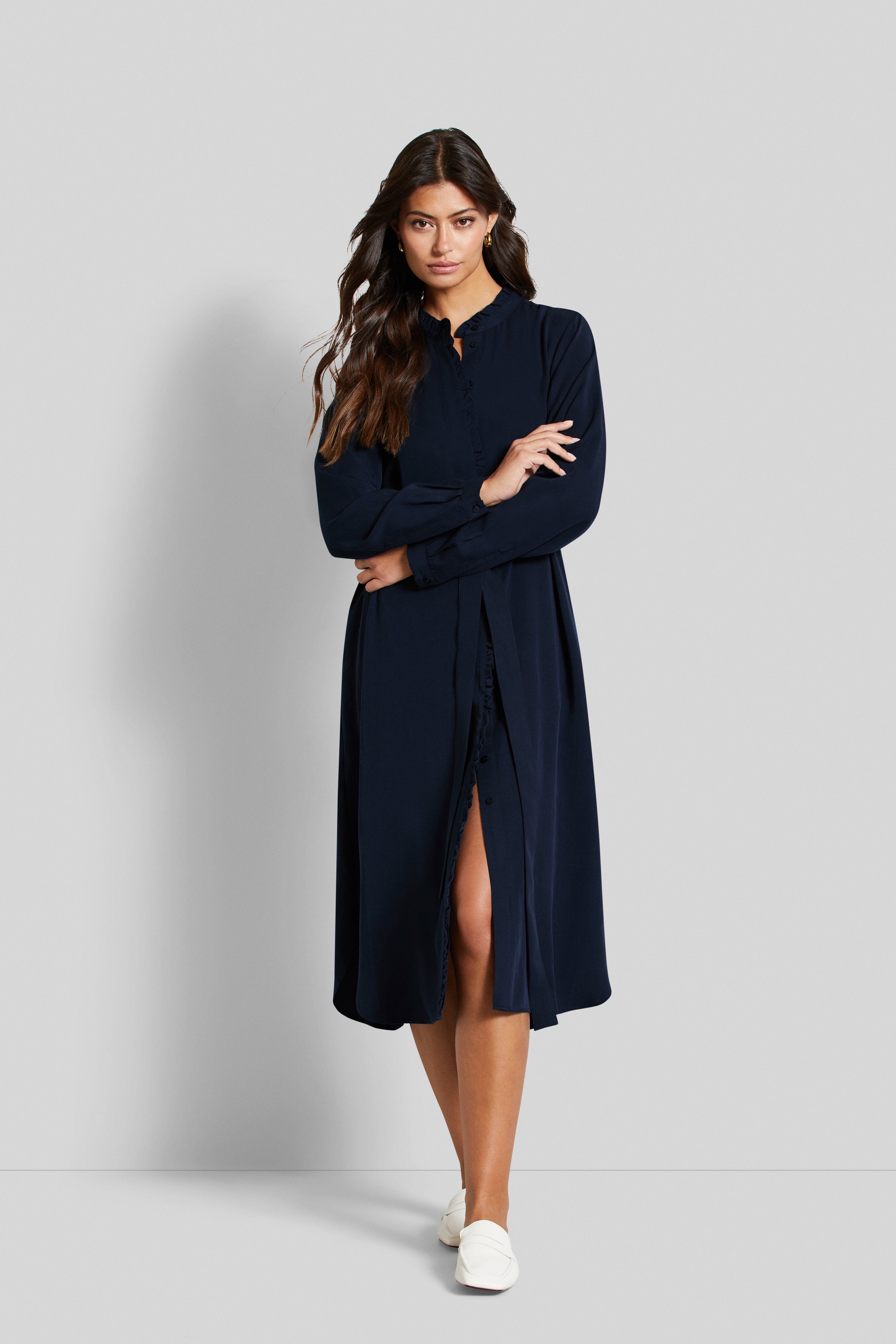 Model in navyblauem bugatti Kleid, Stehkragen, lange Ärmel, subtiler Schlitz, Arme gekreuzt, weiße Schuhe.