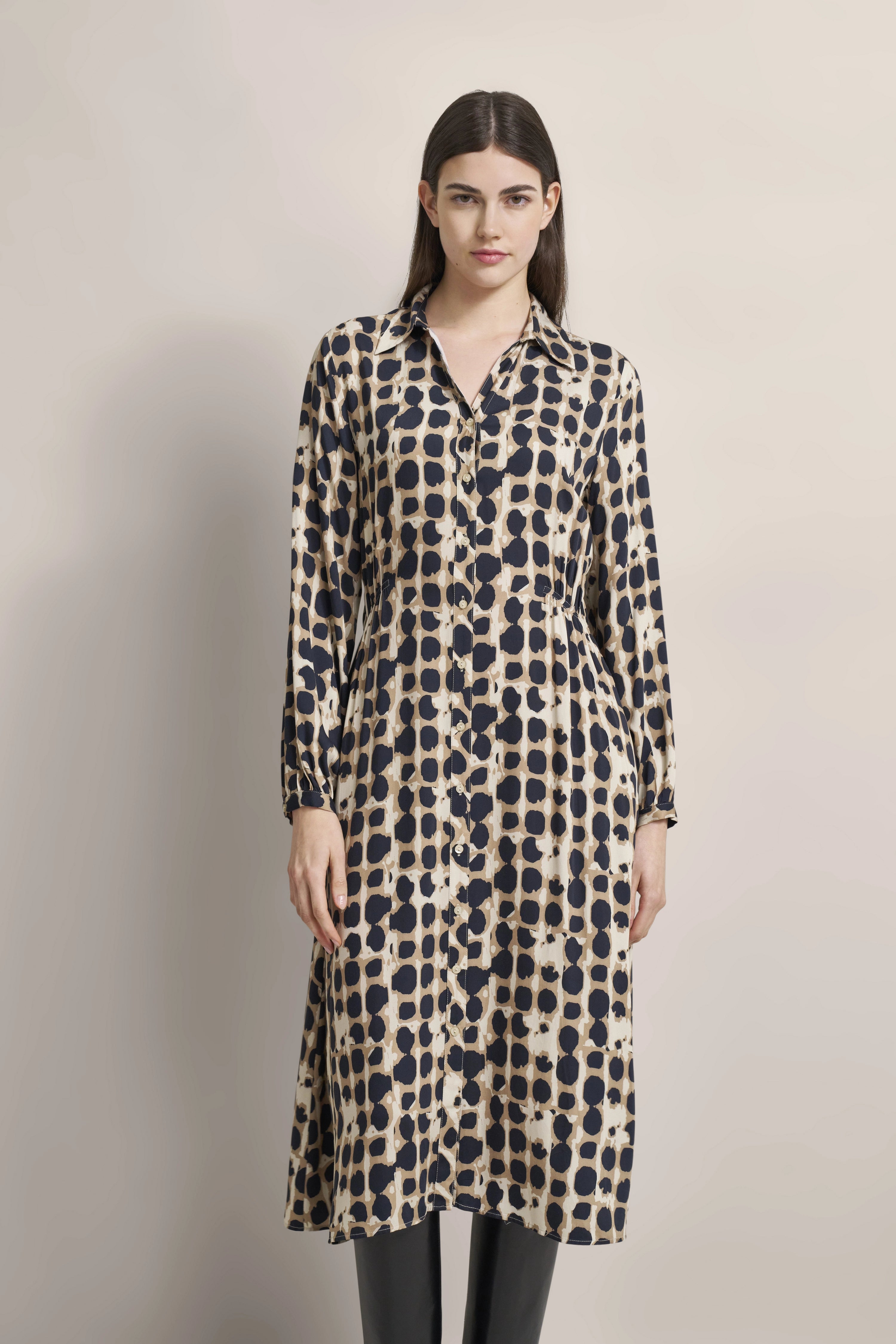 Model wearing long beige and black dress with geometric pattern, relaxed pose.