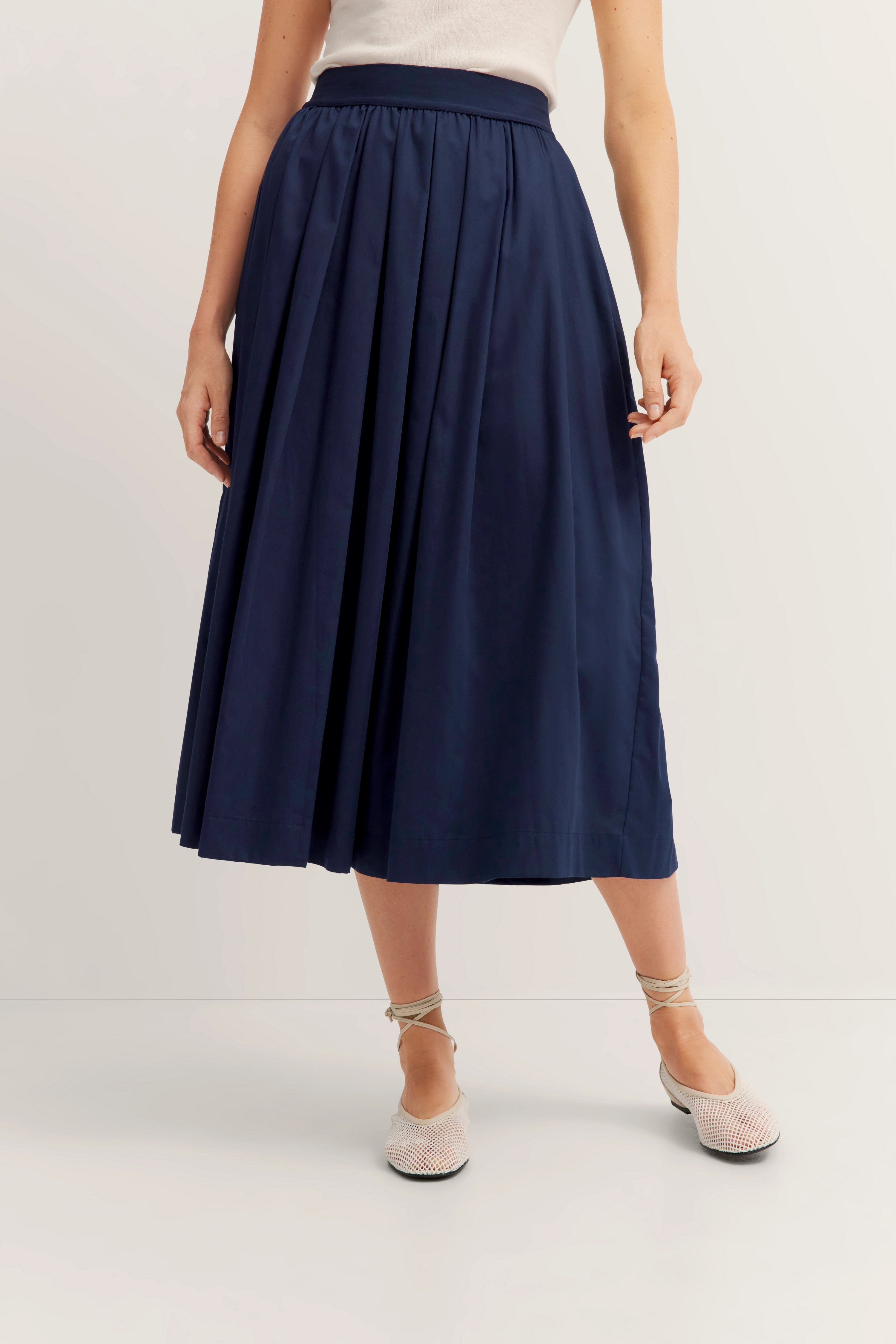 Elegant damesoutfit met blauwe midirok en wit topje, model staat ontspannen.