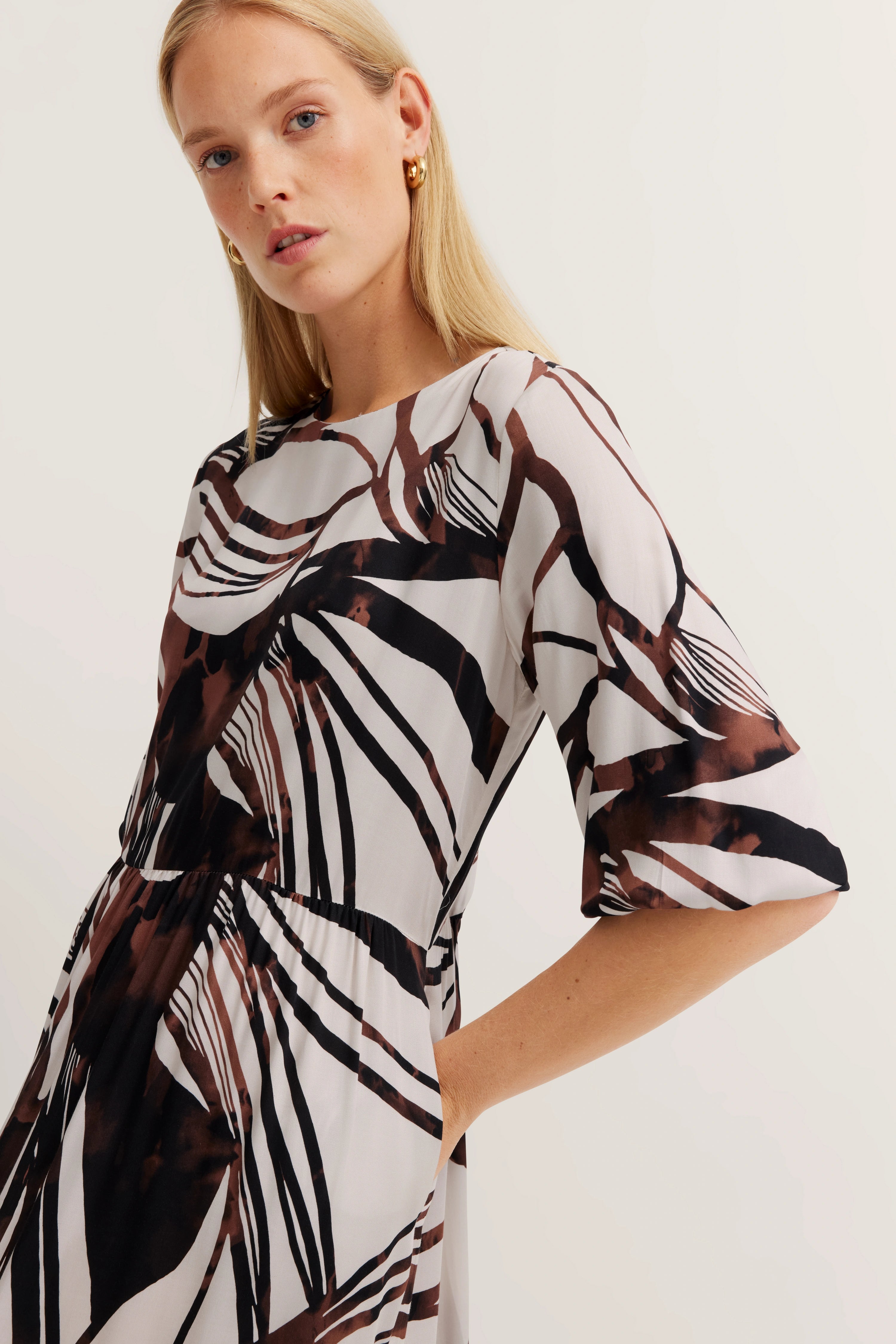 Model in bugatti-jurk met abstract bruin-wit patroon, ontspannen pose.