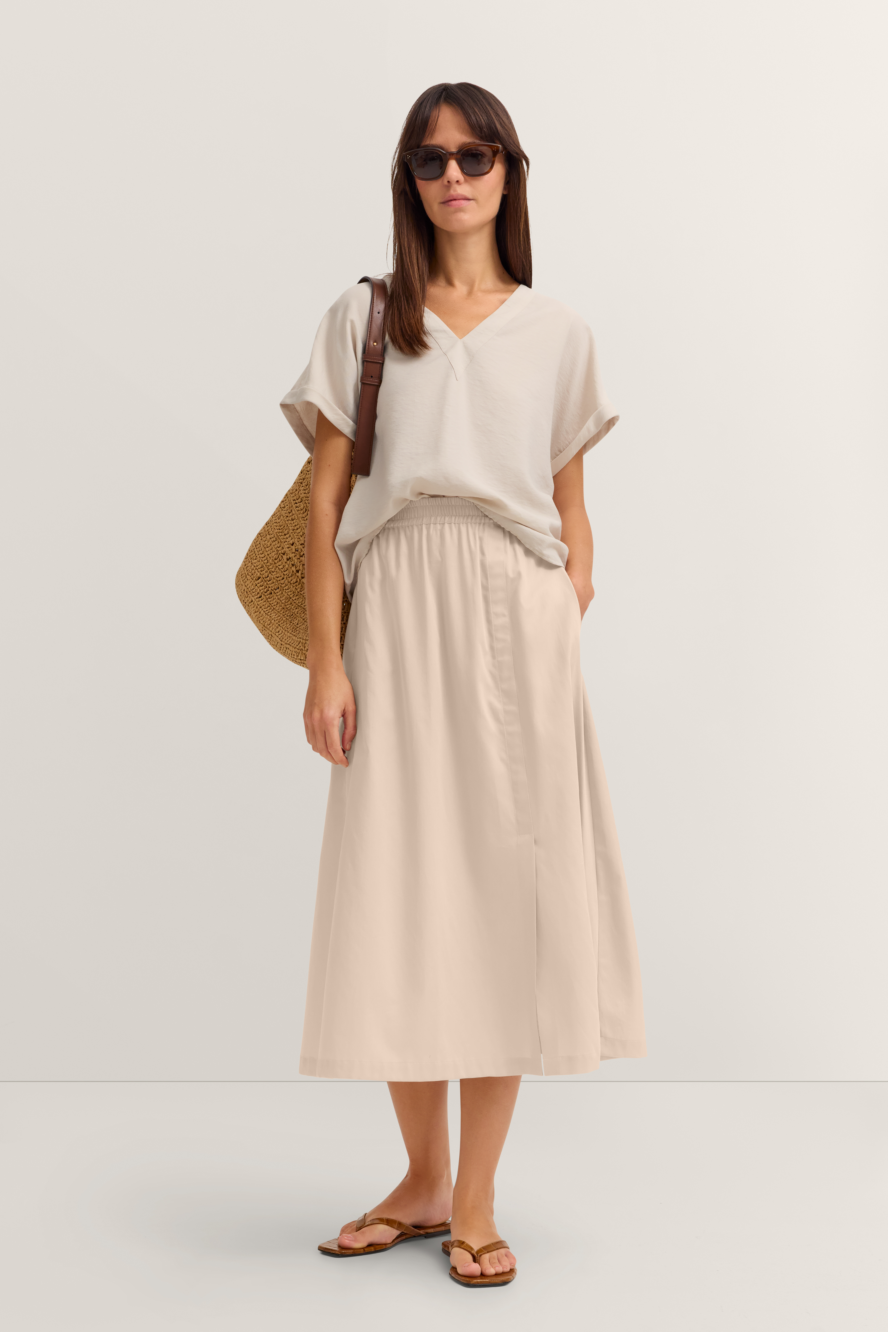 Model in beige T-Shirt und Rock von bugatti, mit Tasche und Sandalen, lässiges Stehen.