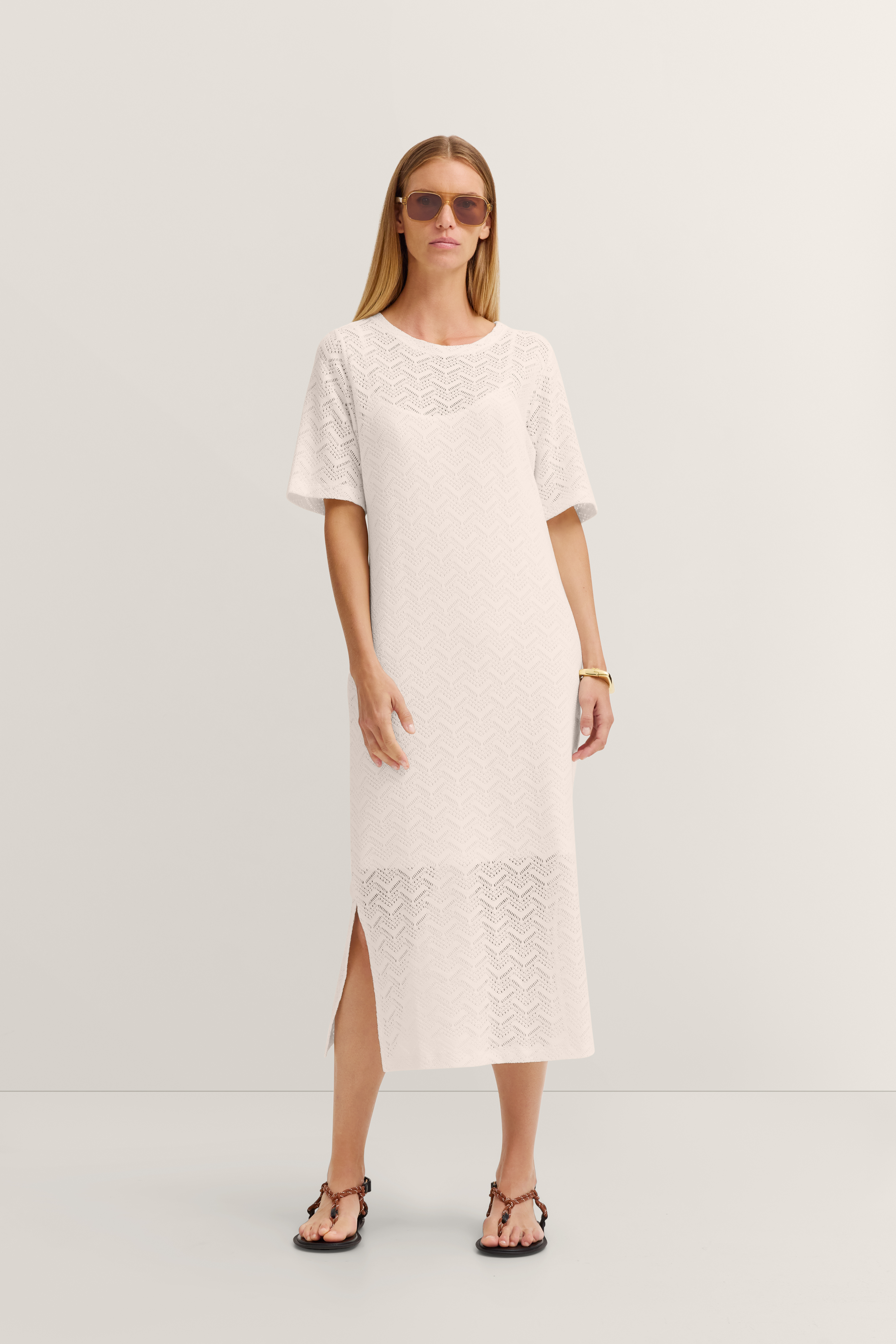 Weißes Spitzenkleid, Model steht entspannt mit Sonnenbrille, Sandalen.