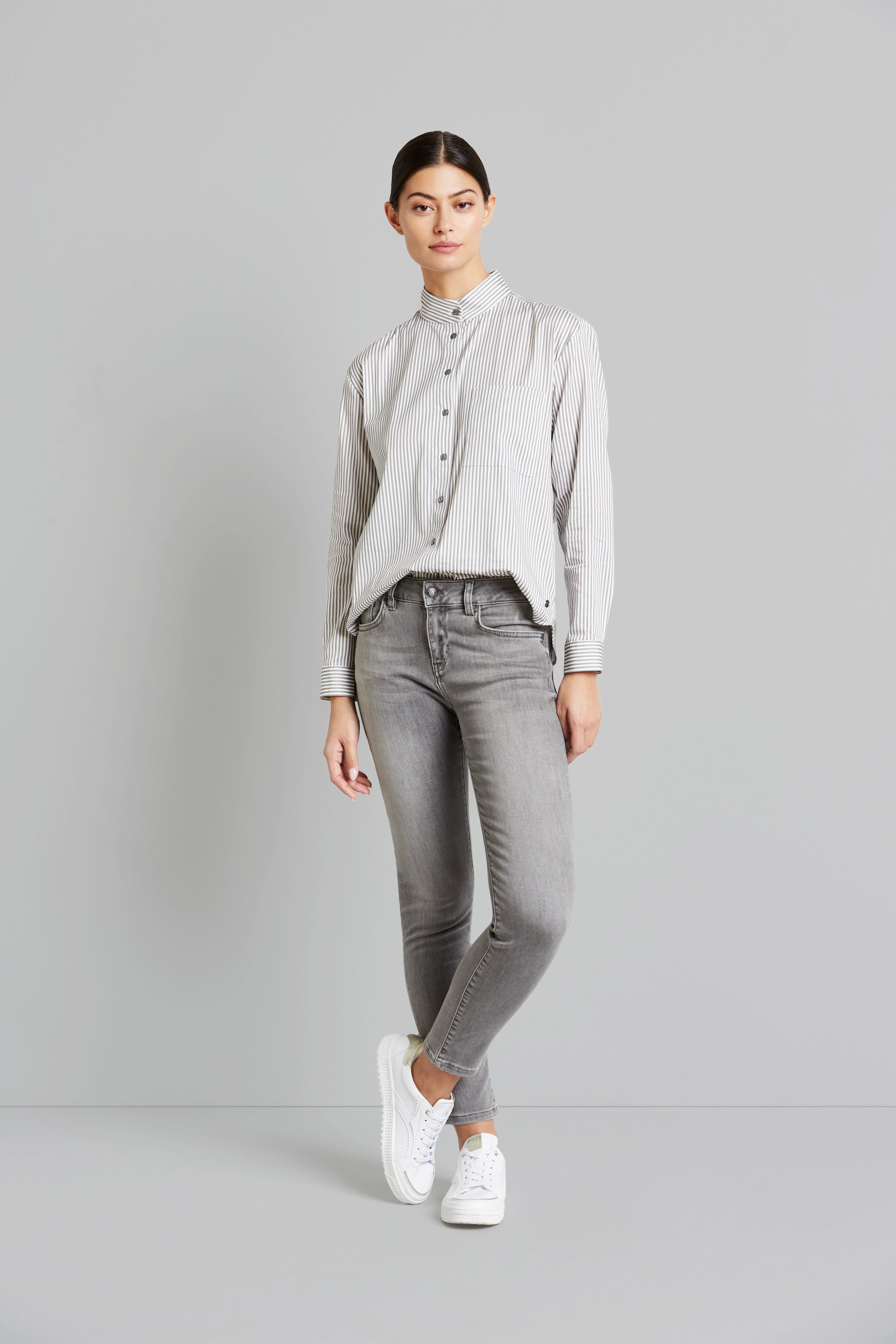 Model in gestreiftem Hemd, grauer Jeans und weißen Sneakers, locker stehende Pose.