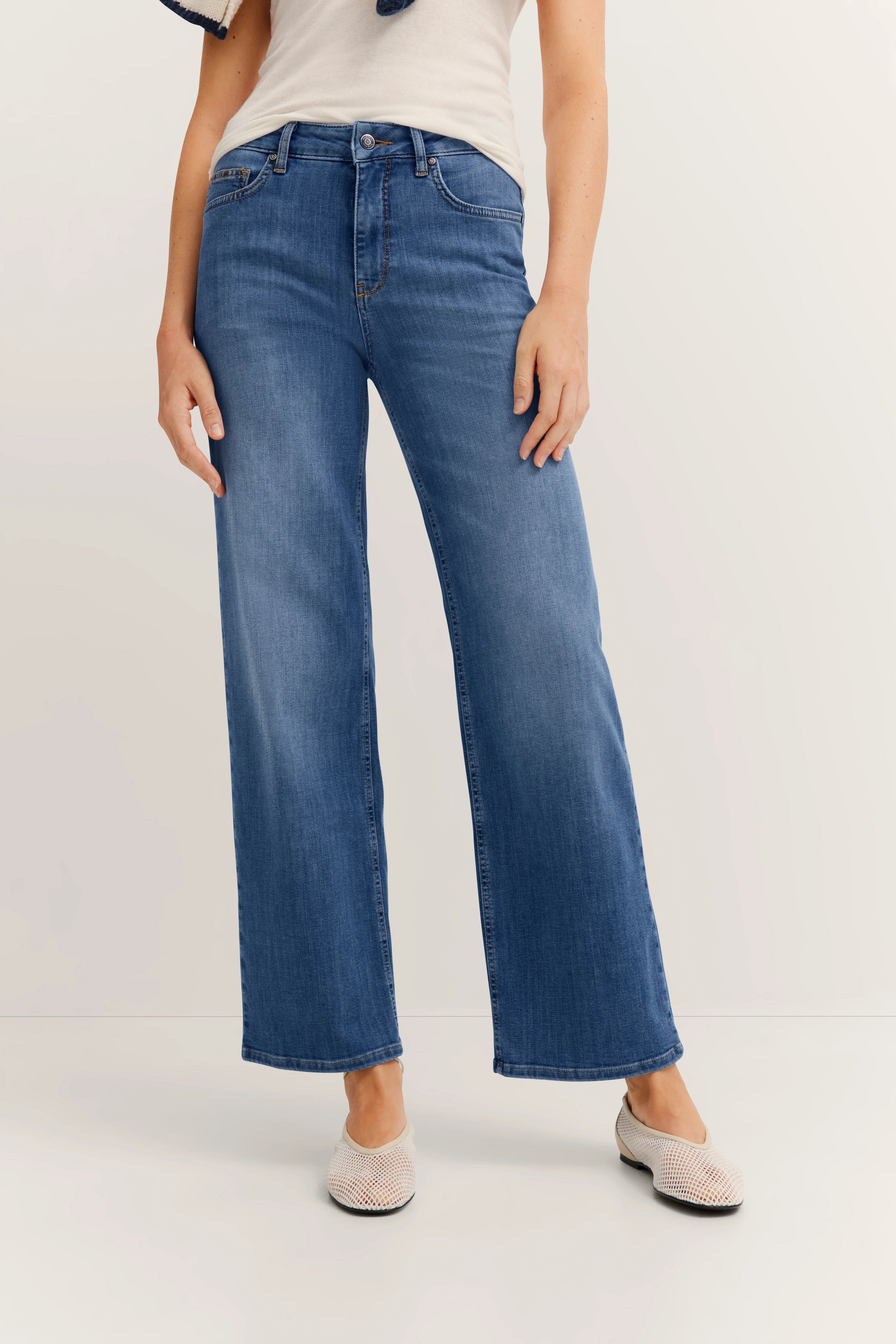 Uitlopende blauwe jeans, casual model, ontspannen pose, gecombineerd met lichte schoenen.