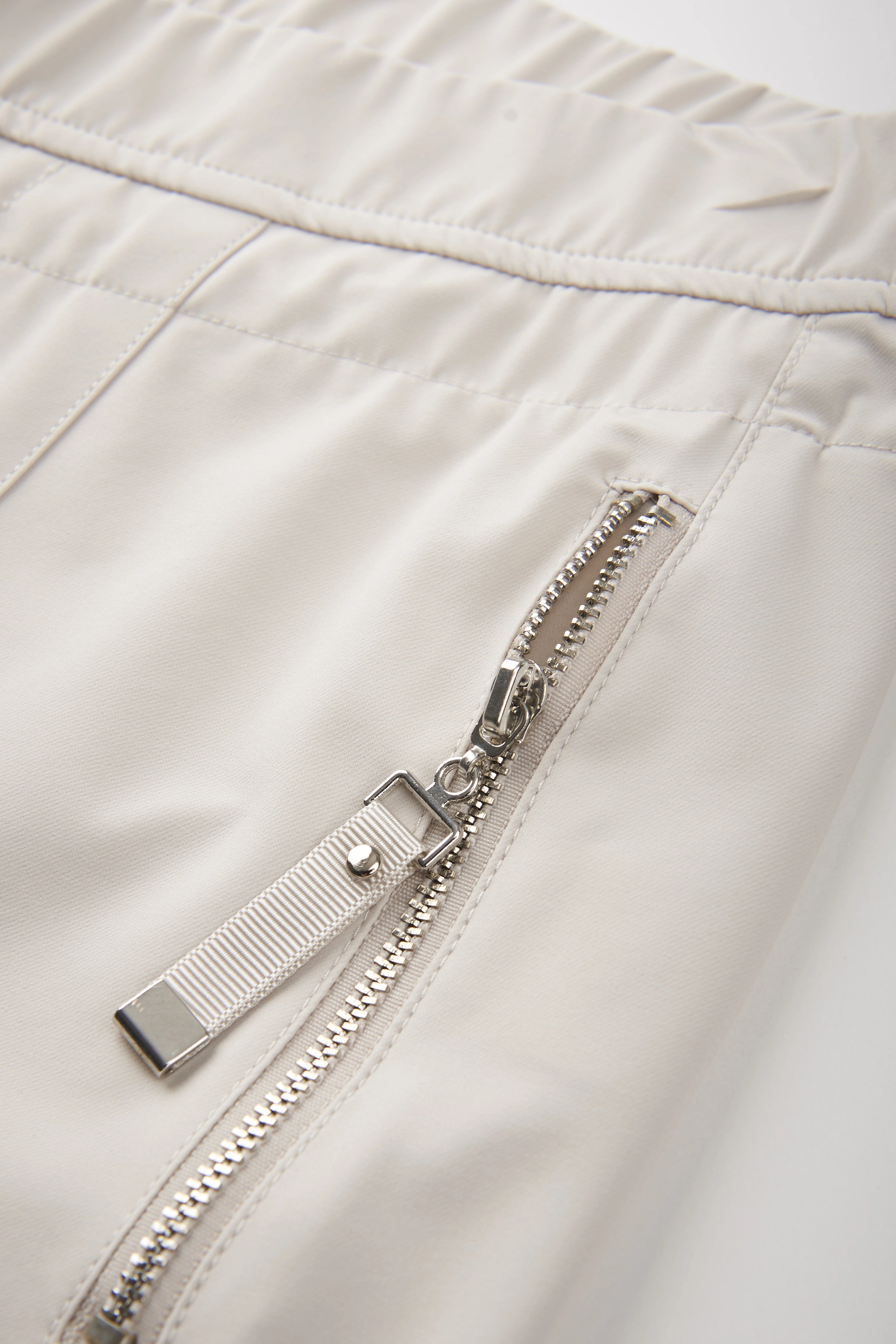 Detailaufnahme einer cremefarbenen bugatti Hose mit silbernem Reißverschluss und elastischem Bund.