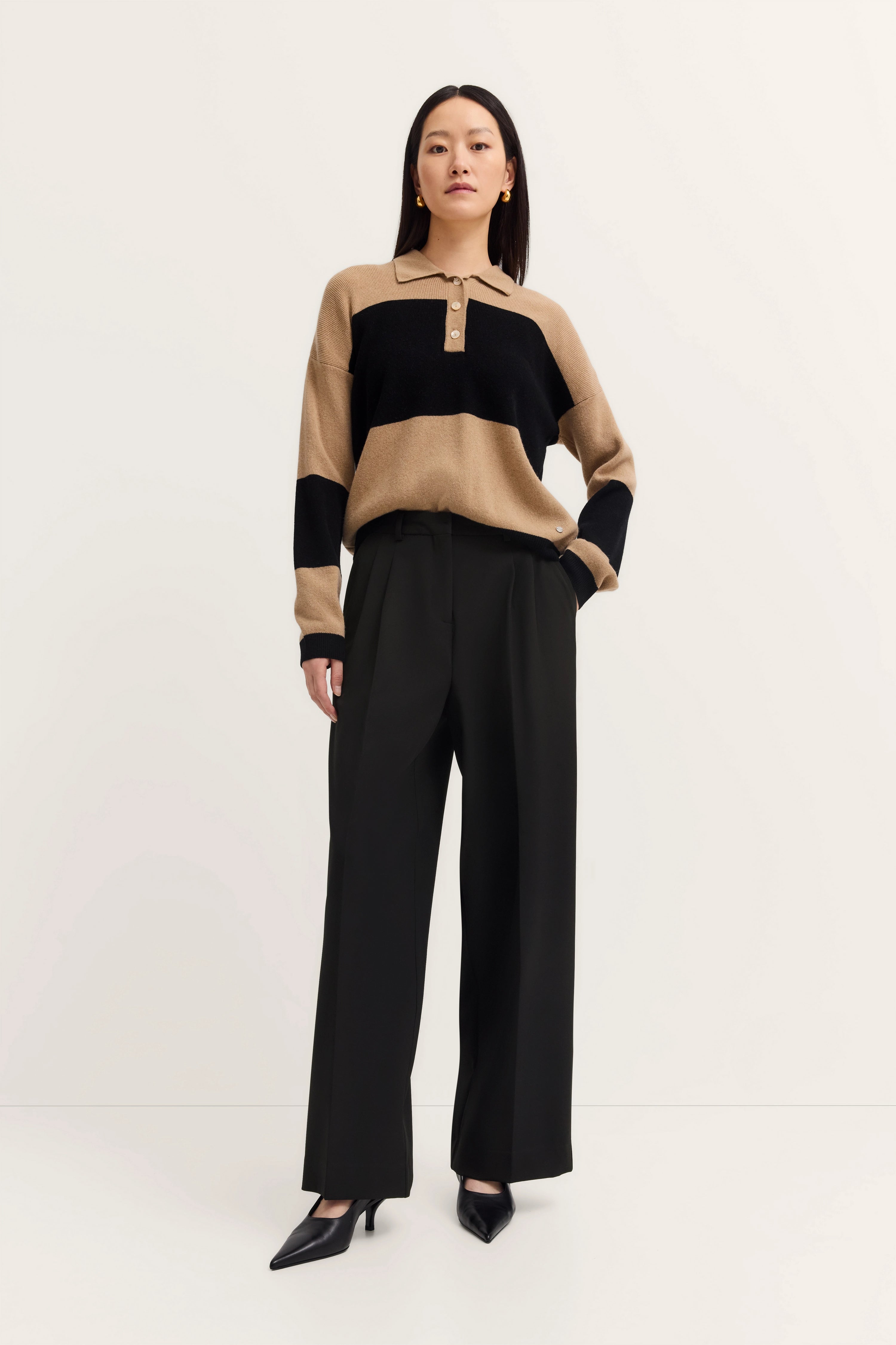 Model in beige-schwarzem Pullover mit Streifen, schwarze Hose, elegante Haltung.