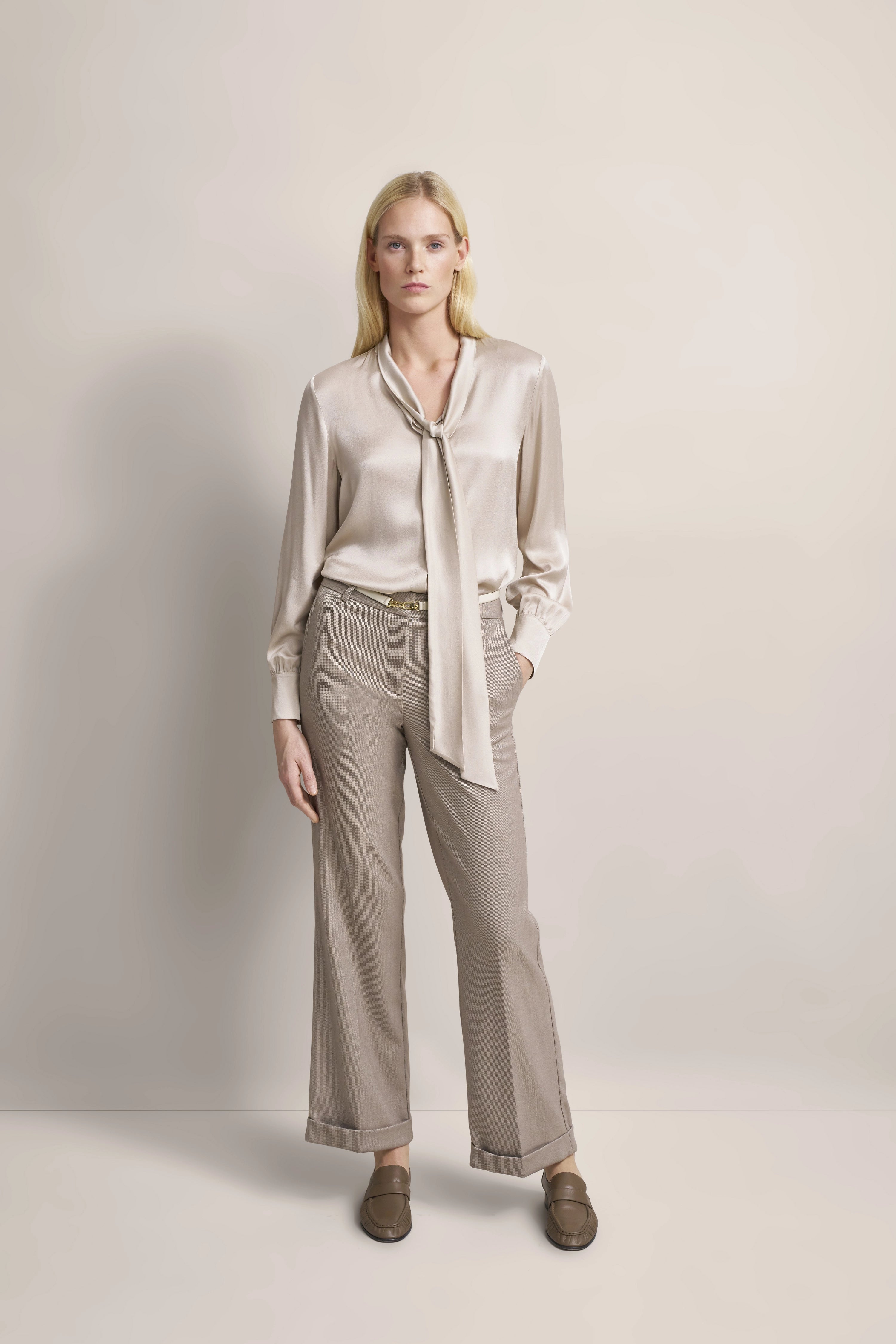 Elegantes Modelbild: Seidenbluse mit Schleife, beige Hose, braune Loafer, entspannte Pose.