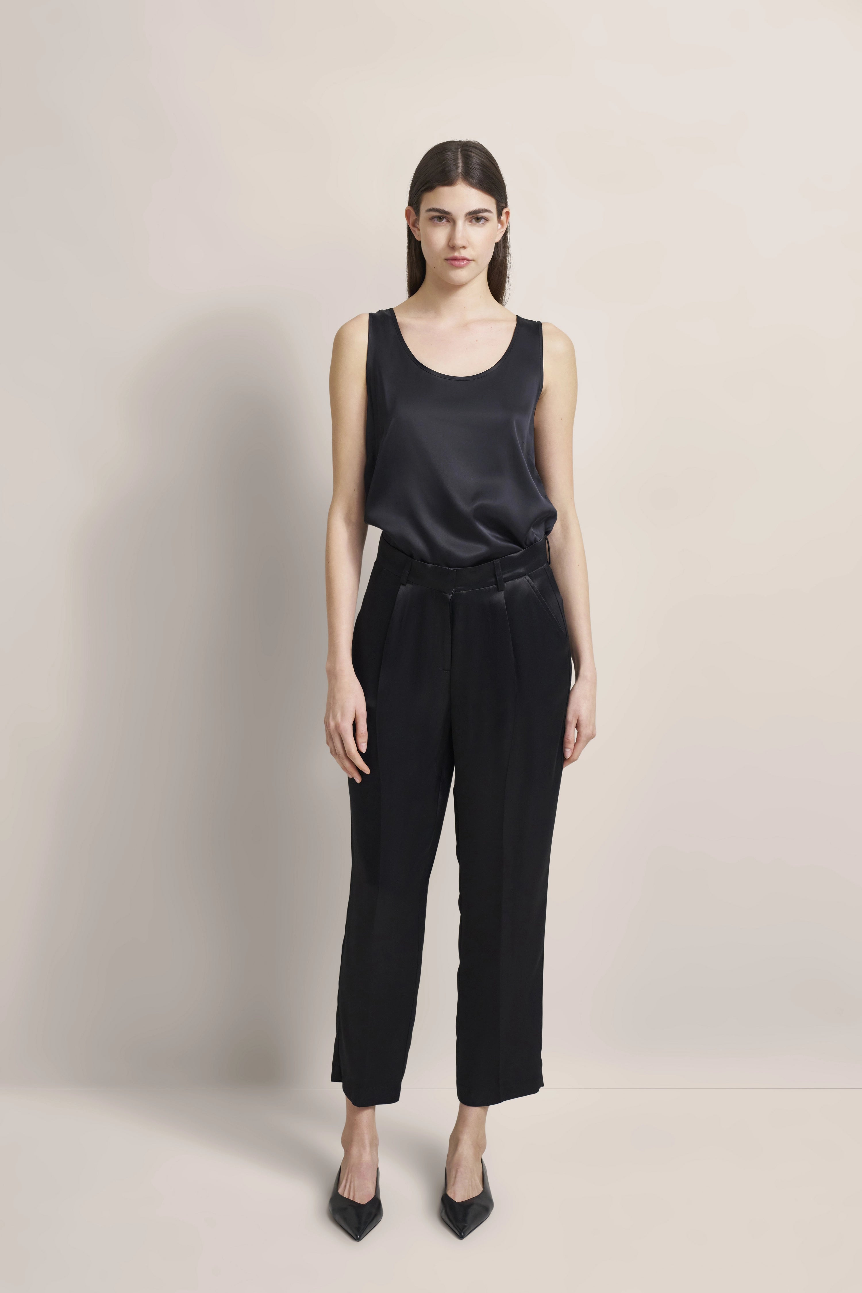 Model trägt schwarzes ärmelloses Top und schwarze Hose, steht aufrecht, neutraler Ausdruck.